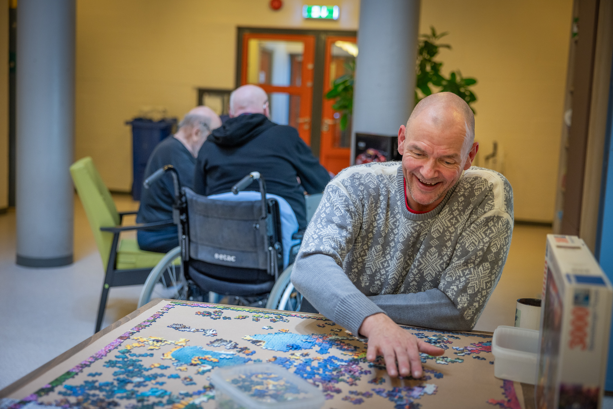 Skallet, middelaldrende og smilende mann med lys grå strikkegenser sitter og pusler puslespill ved et bord. I bakgrunnen sitter to andre menn rundt et bord.