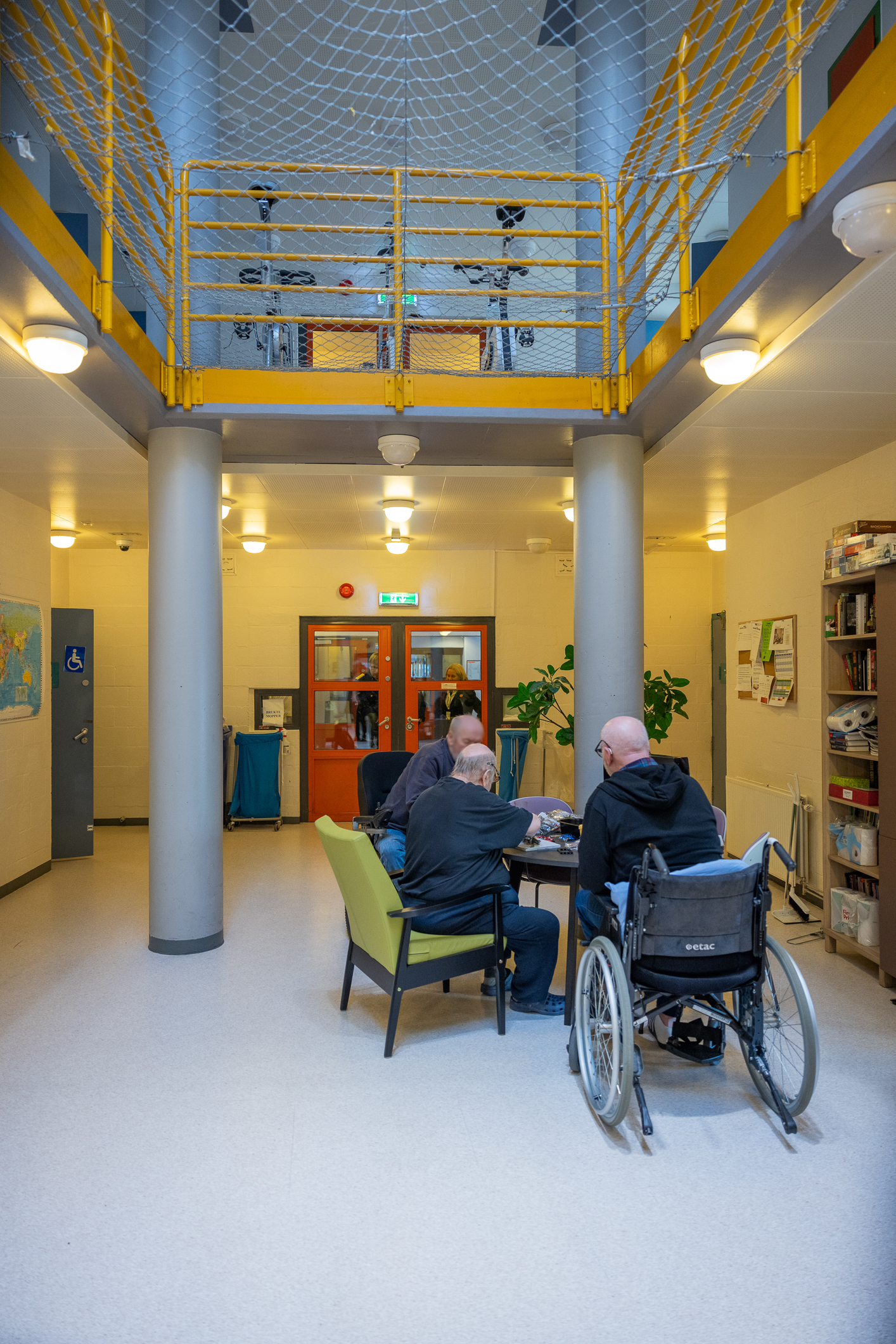 Tre menn sitter oppslukt i aktivitet rundt et bord, med ryggen til rundt et bord. Gul vegg og rød dør. Rommet er stort og er åpent opp til etasjen over der man ser tre treningssykler. Et stort nett skal hindre noen å falle ned på gulvet i etasjen som mennene  sitter.