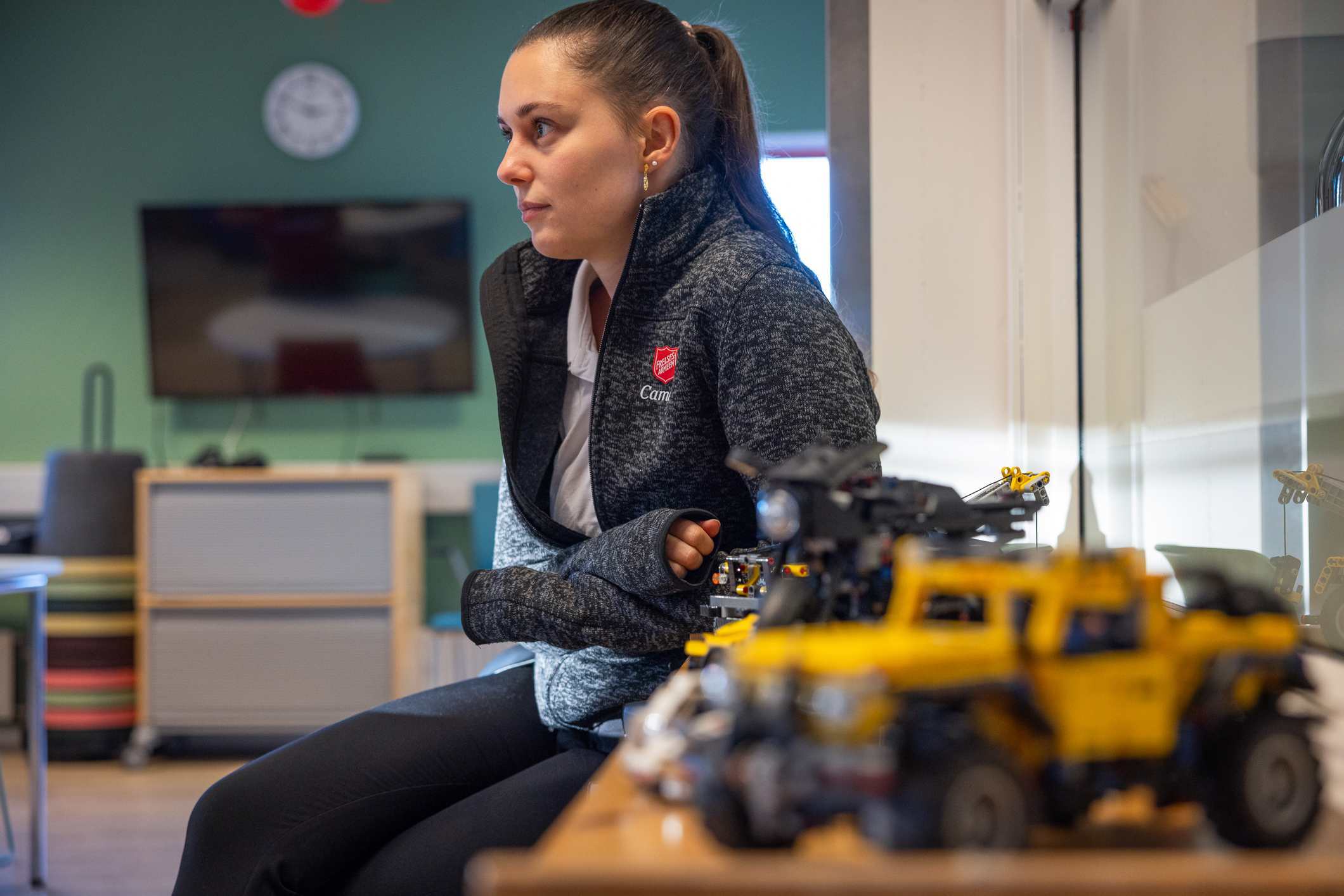 Kvinne med mørkt hår og hestehale som sitter ved et bord i et aktivitetsrom. Hun har Frelsesarmeens arbeidstøy på. En grå fleecejakke med rødt skjold på brystet. Et par lekebiler står på bordet i forgrunnen