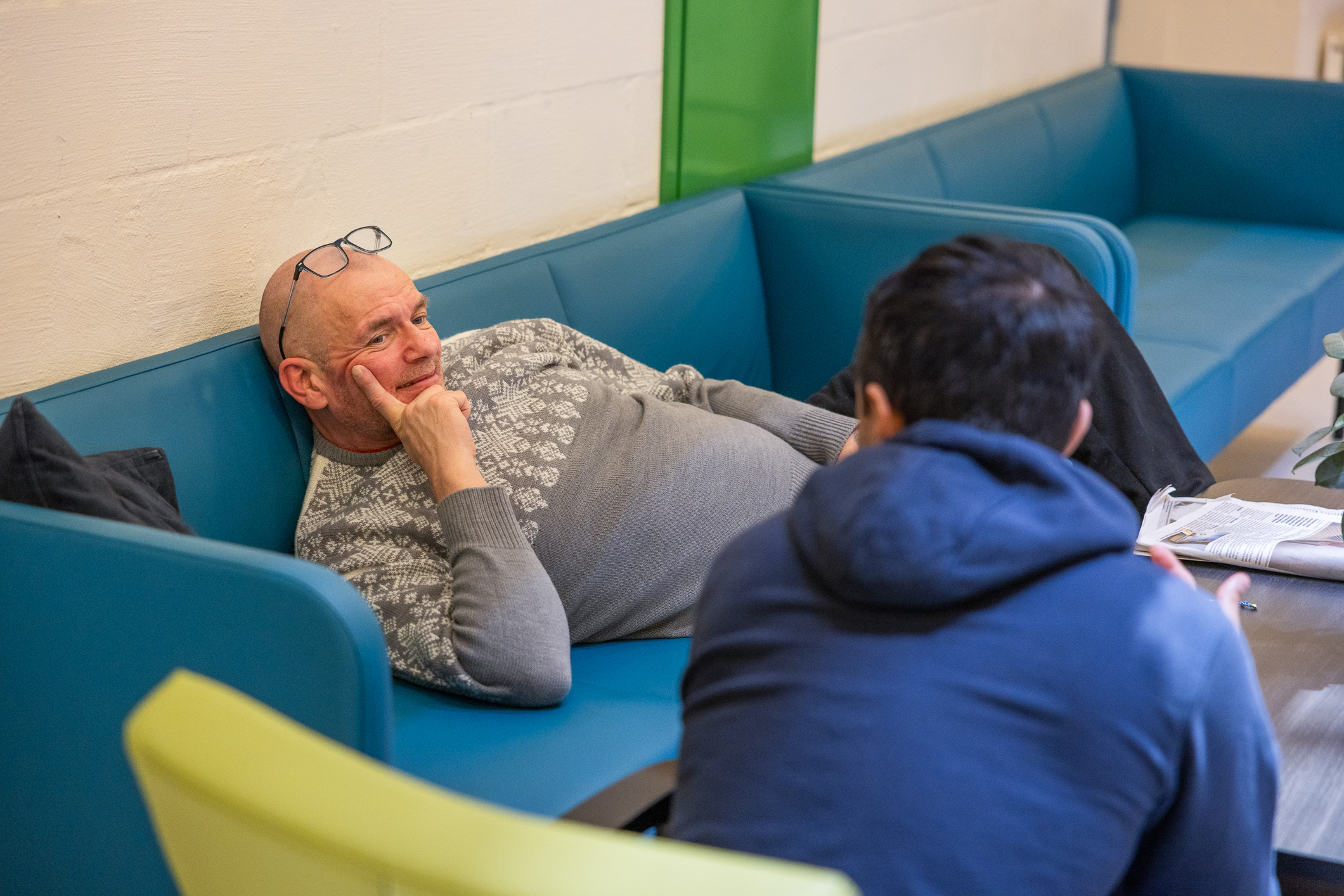 Middelaldrende, skallet mann med lys grå strikkegenser ligger og slapper av på en blå sofa. Han snakker med en yngre mann med mørkt hår som sitter med ryggen til.