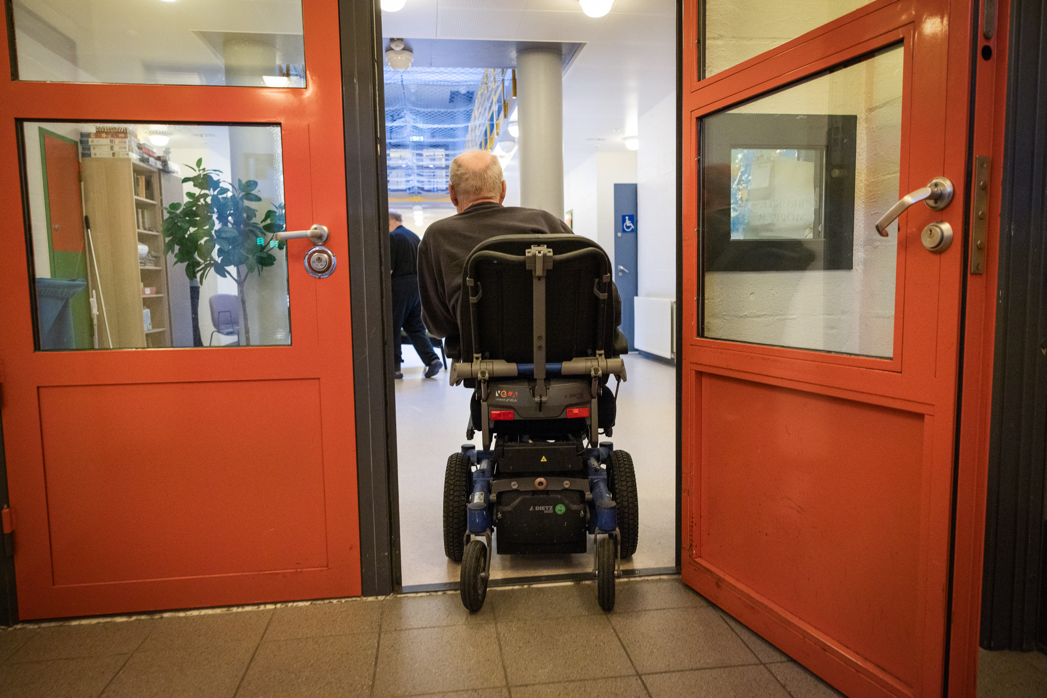 Foto: Sett bakfra, kjører en rullestolbruker gjennom to røde dører på en avdeling i et fengsel.