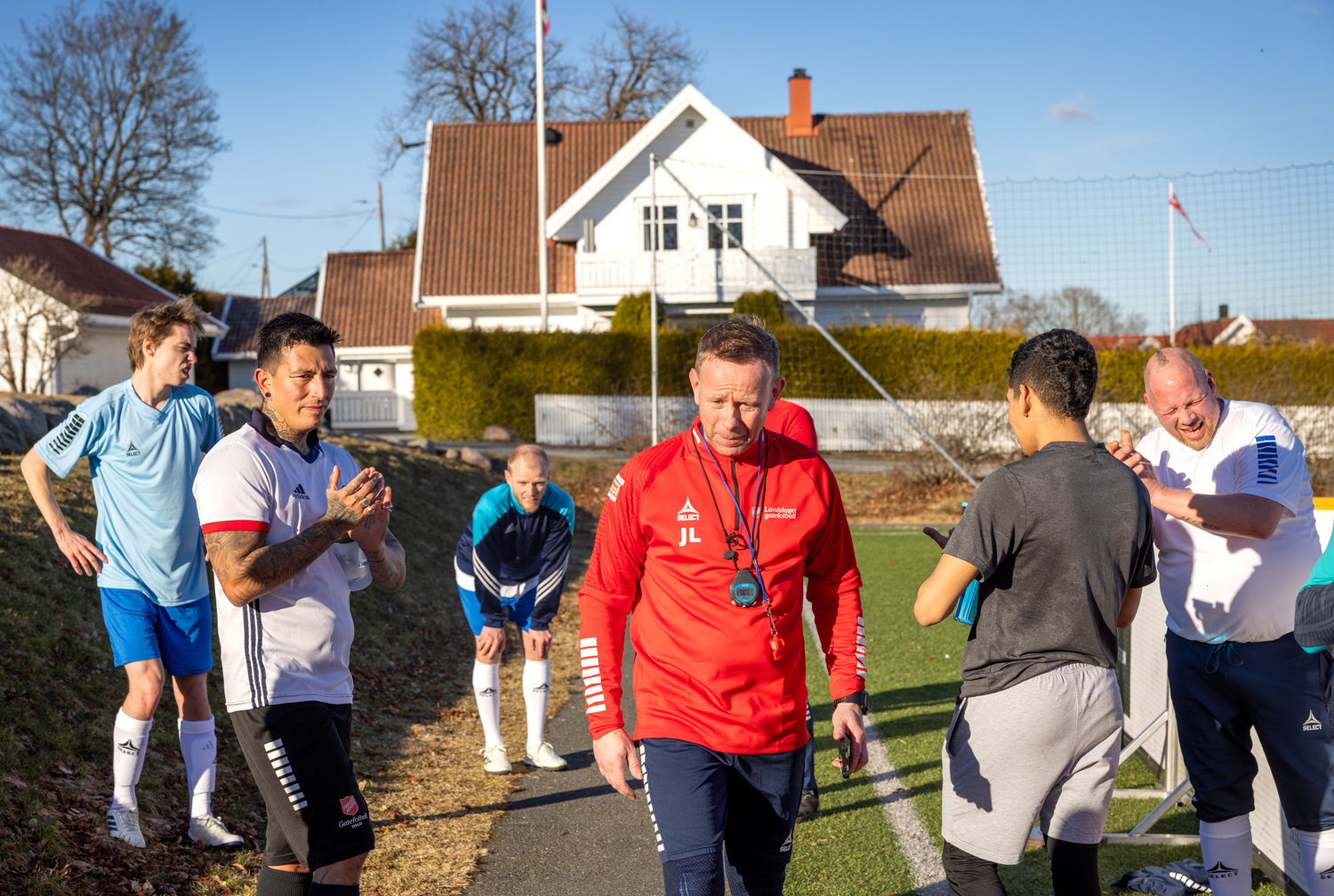 Foto: Mann i rød treningsjakke går gjennom en gruppe med forballspillere like utenfor banen. I bakgrunnen sees et hvitt hus.