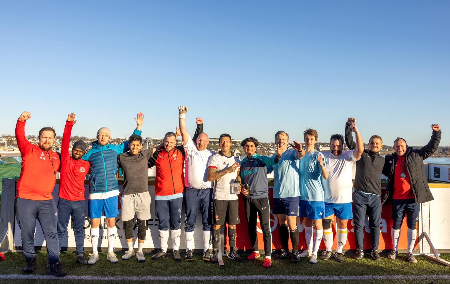 Foto: 13 menn står oppstilt på rekke i en fotballbinge. De holder hendene opp.