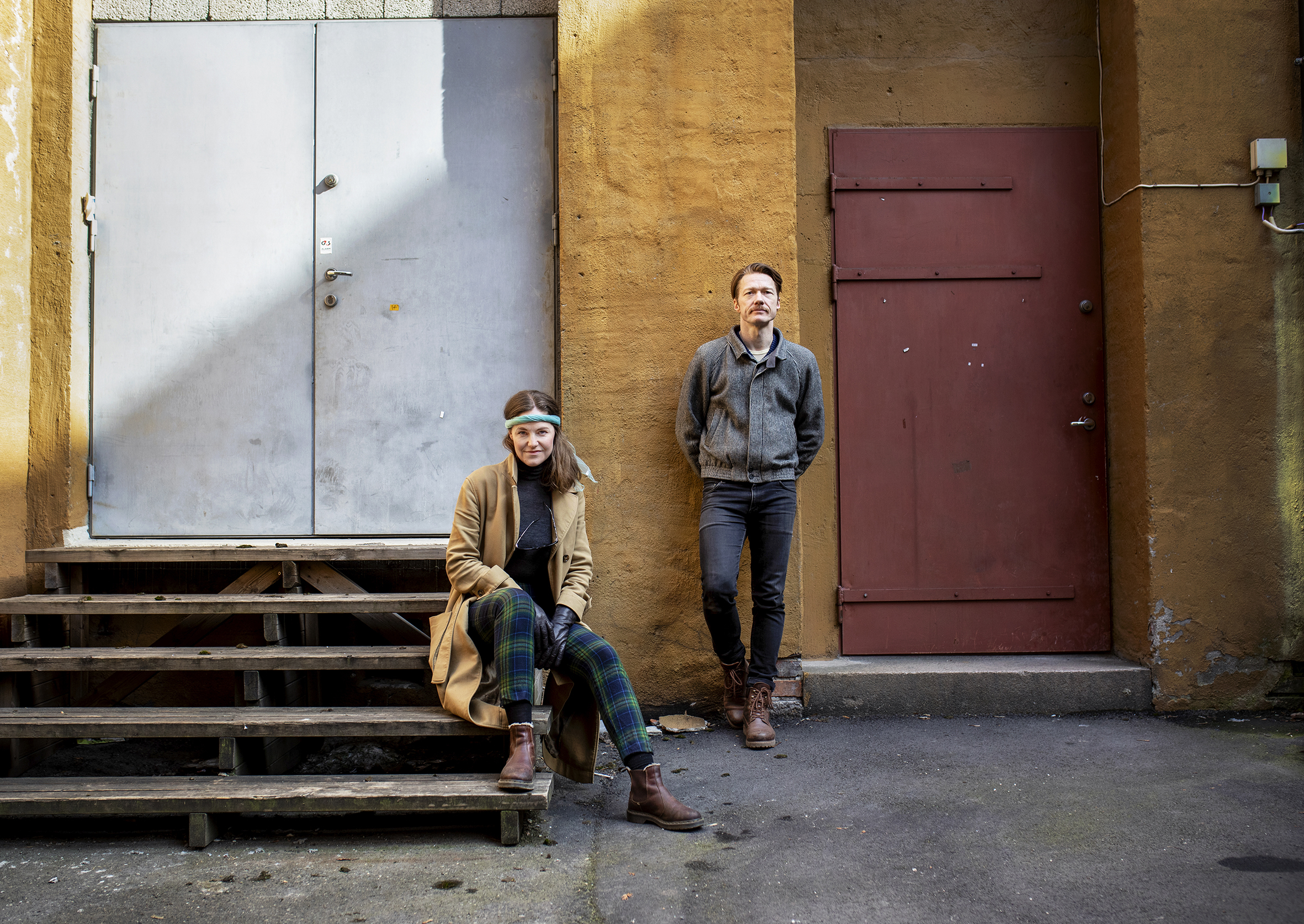 Mari Sandvær Kreken sitter på en tretrapp som leder opp mot en grå ståldør. Tor Egil Kreken står lent mot en gul murvegg ved siden av. De ser i kameraet.