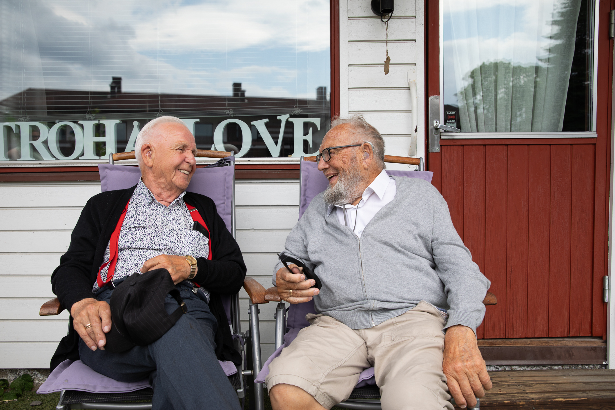 To gamle menn sitter i hver sin hagestol utenfor en inngangsdør og et vindu.