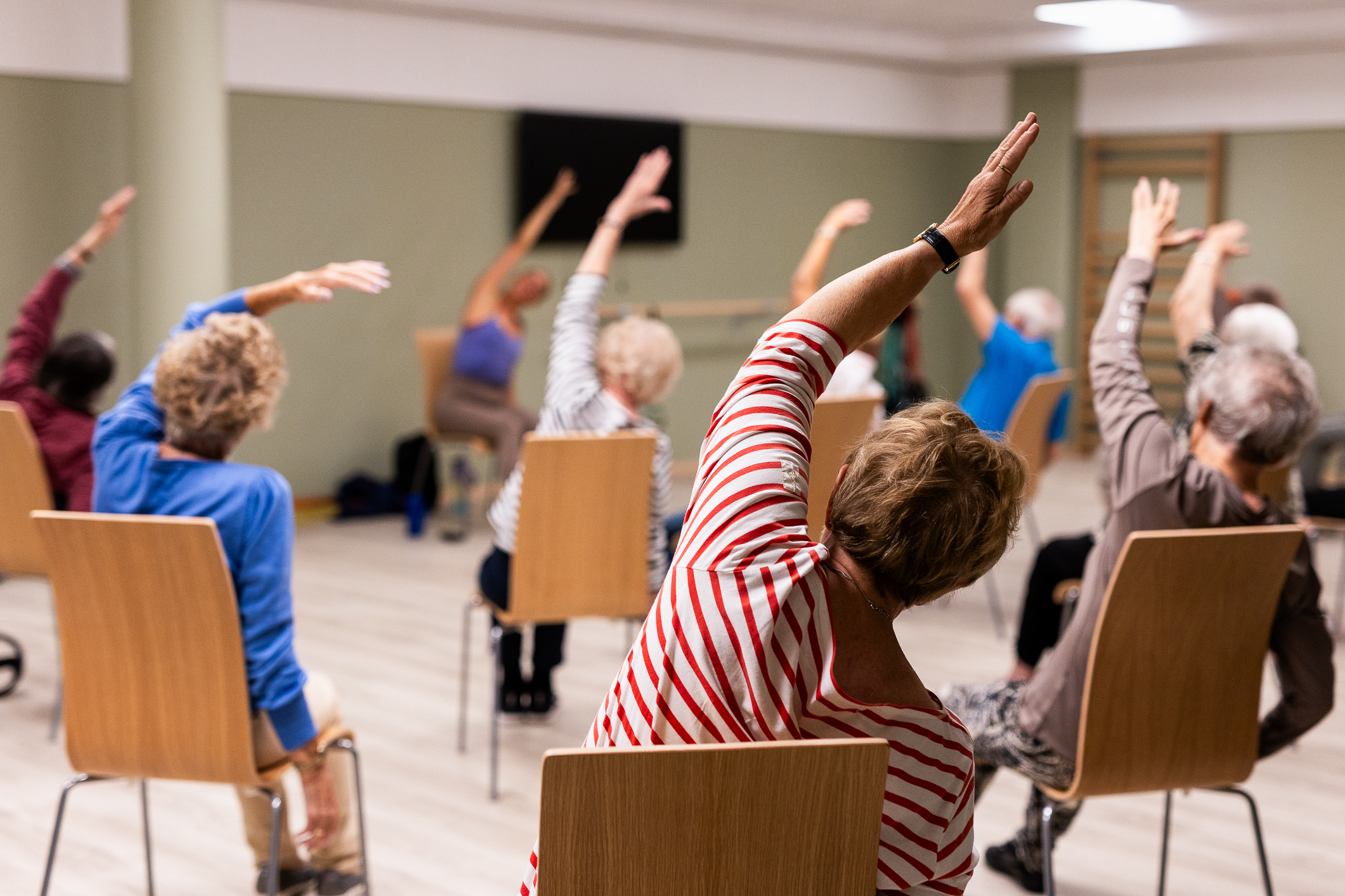 Eldre som sitter på stoler og bøyer og tøyer. Alle sitter med ryggen til, men vendt mot en instruktør.
