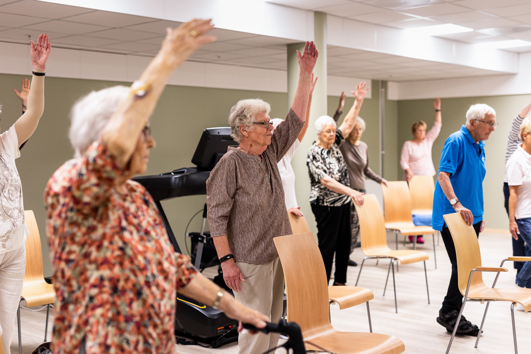 Flere eldre som står bak stolene sine og strekker ene armen opp.