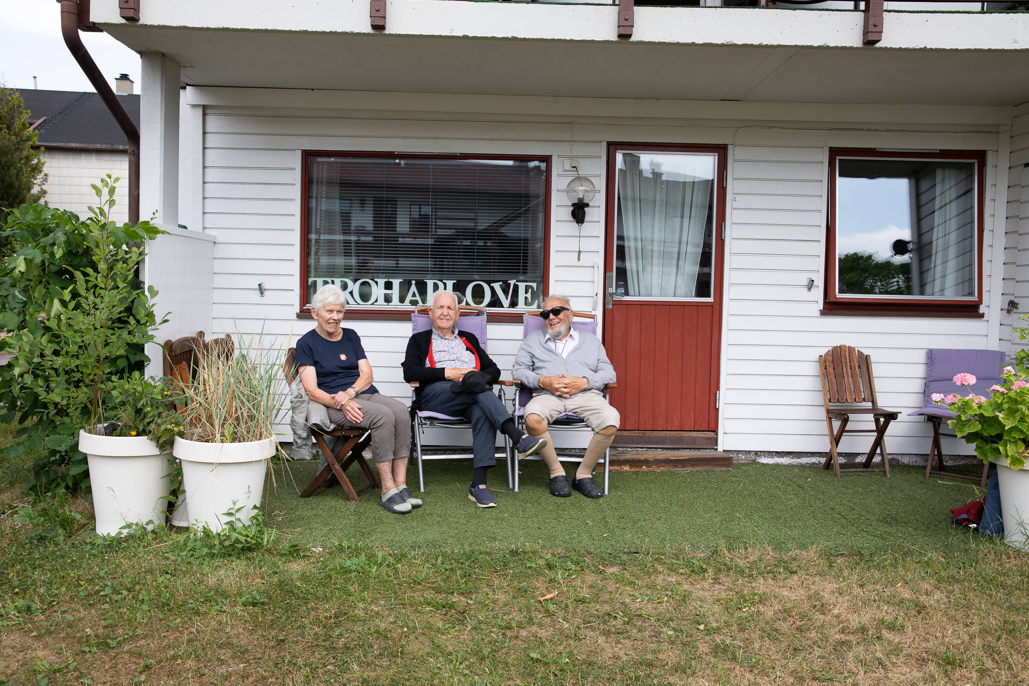Kvinne og to menn – alle pensjonister – sitter på rekke foran en husvegg.