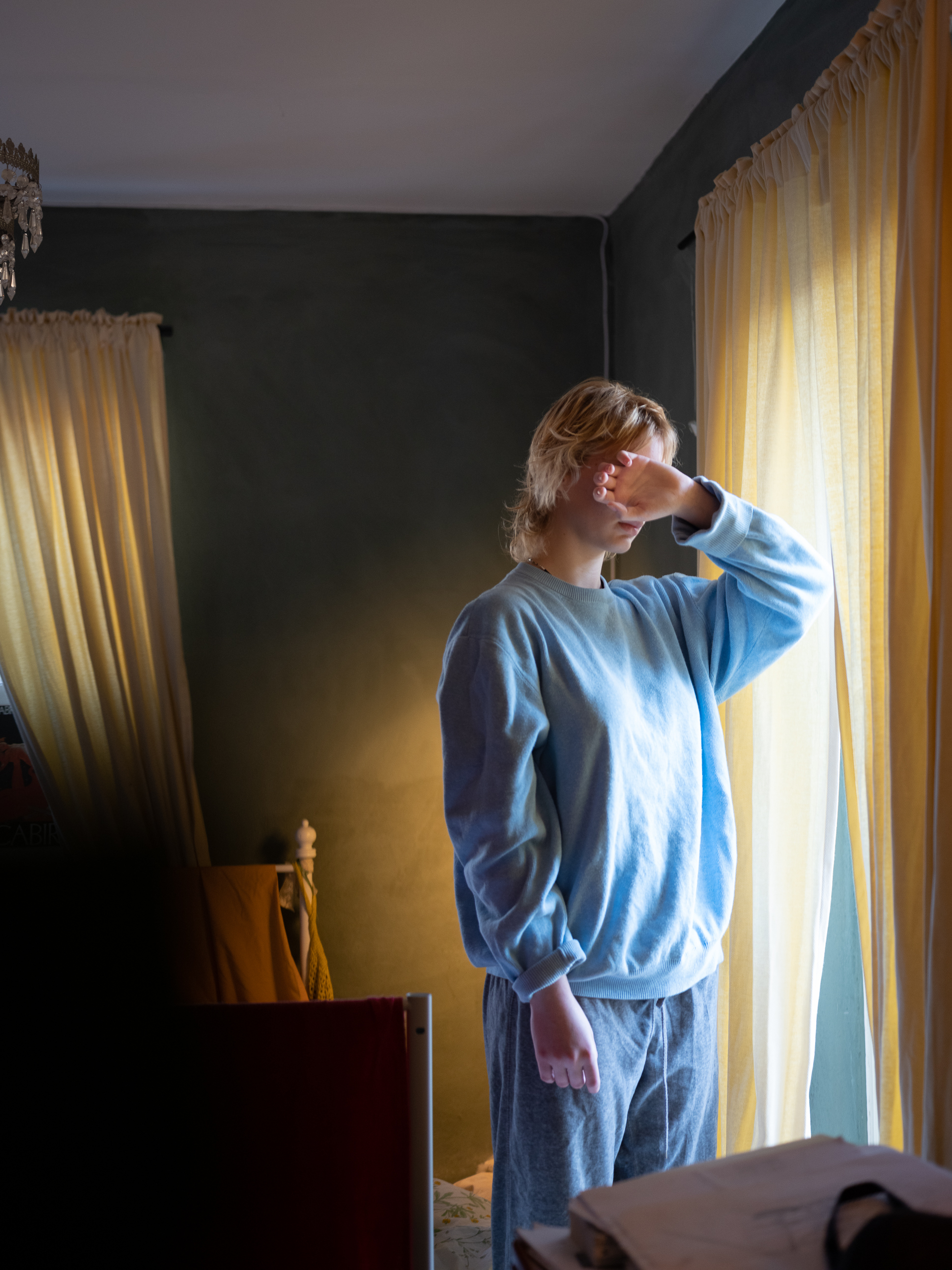 En jente skjuler ansiktet med armen og står vendt mot et vindu