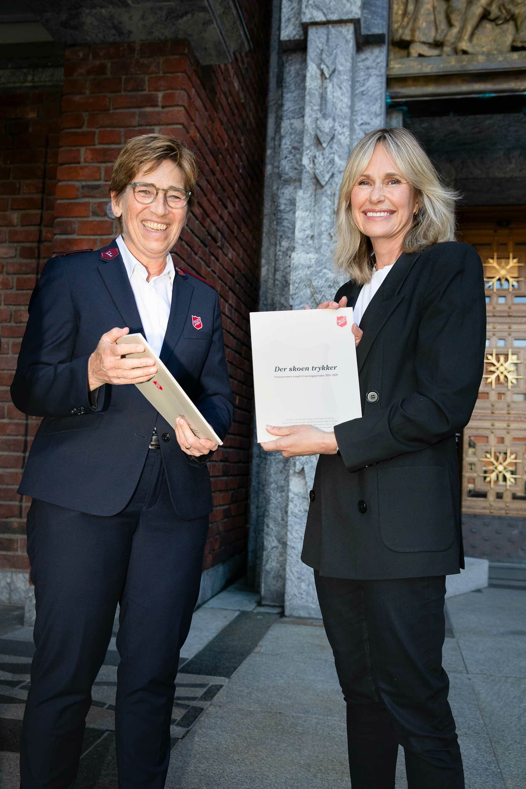 To dresskledde kvinner smiler foran et inngangsparti. Den ene holder en rapport, den andre har Frelsesarmeens skjold på jakkeslaget.