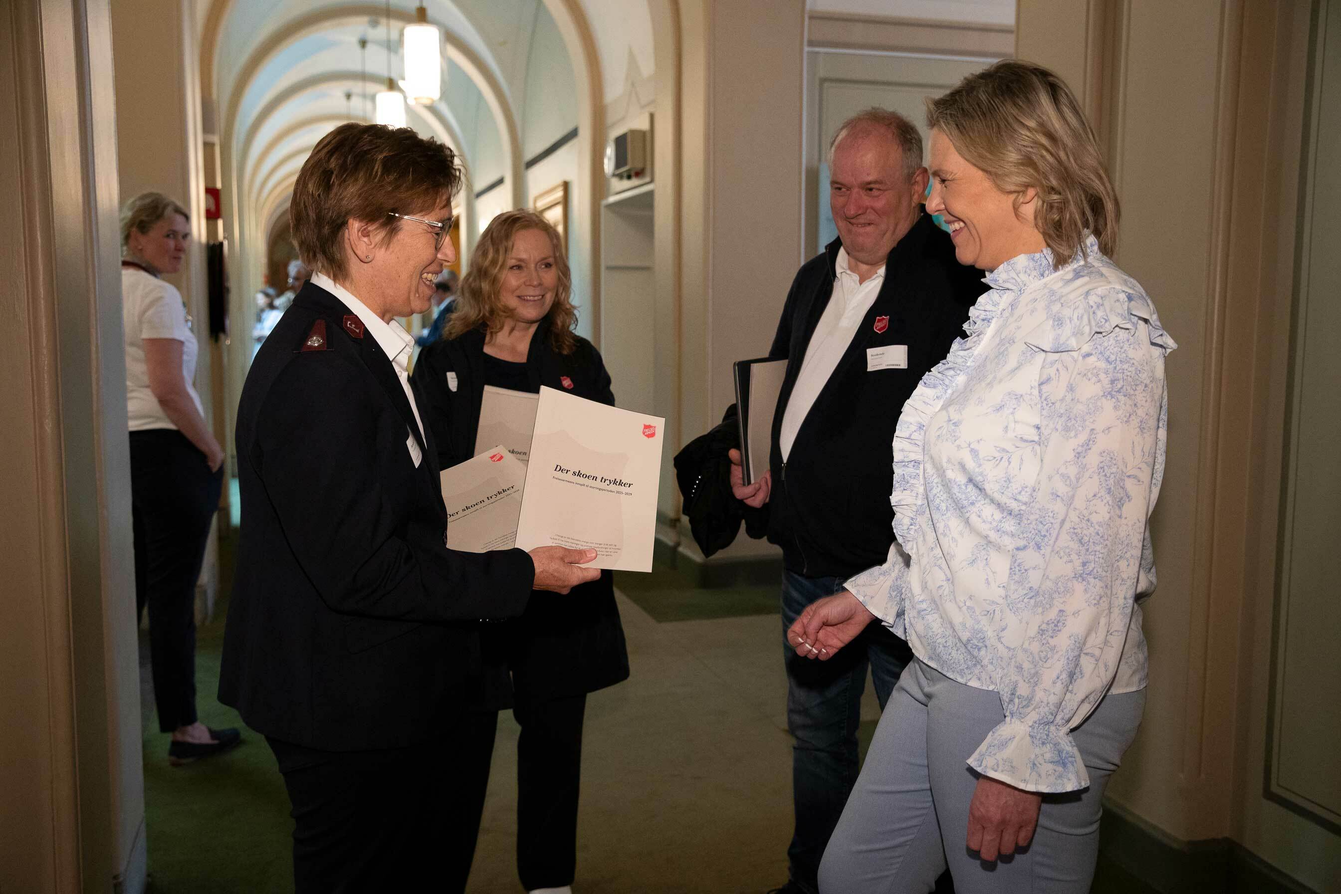 To kvinner og en mann med Frelsesarmeens kjole på jakkeslaget overrekker en rapport til en kvinne med lys bluse.