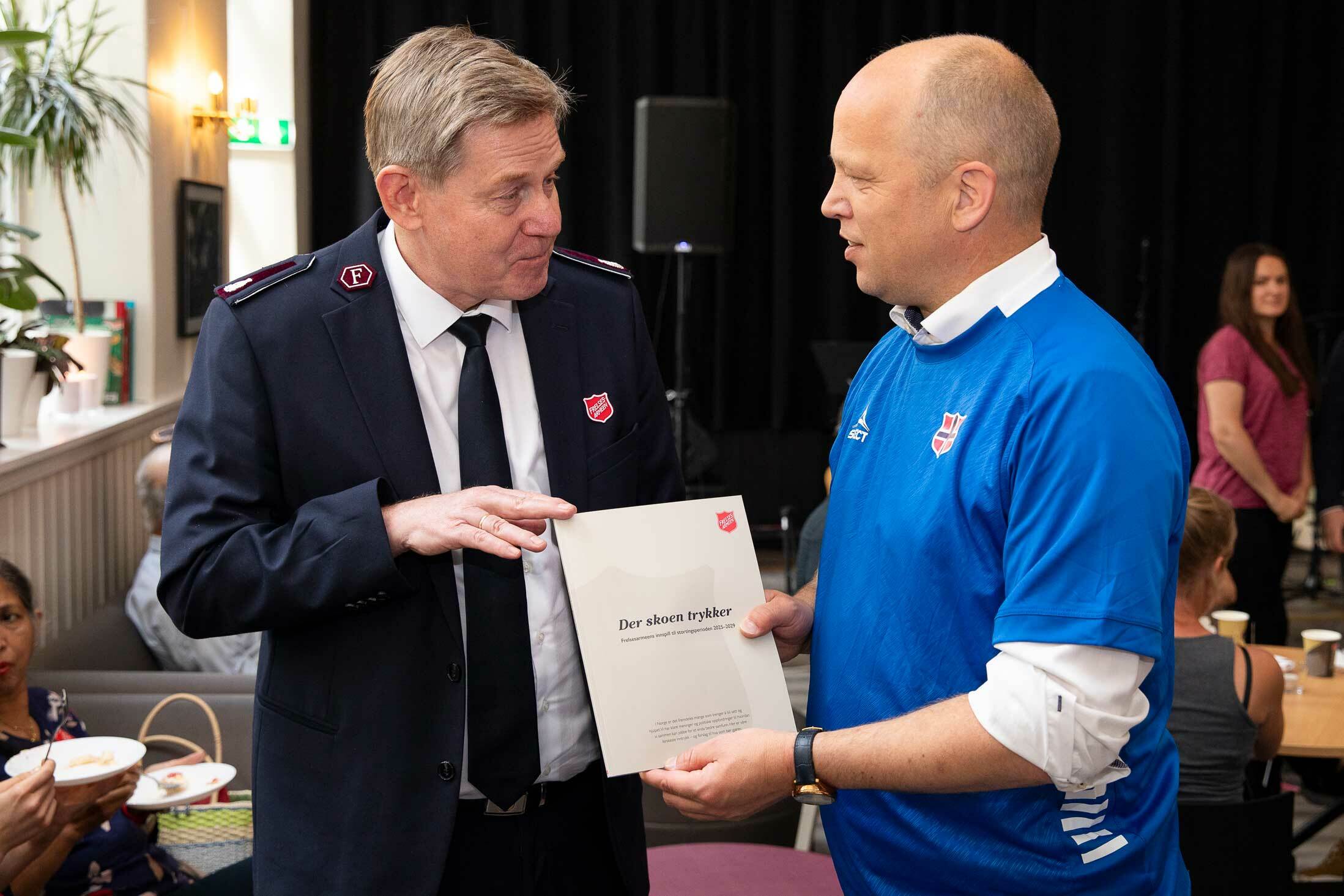 To menn holder en rapport mellom seg. Mannen til venstre har  Frelsesarmeens skjold på brystet. og mannen til høyre har gatefotballens landslagsdrakt på seg.