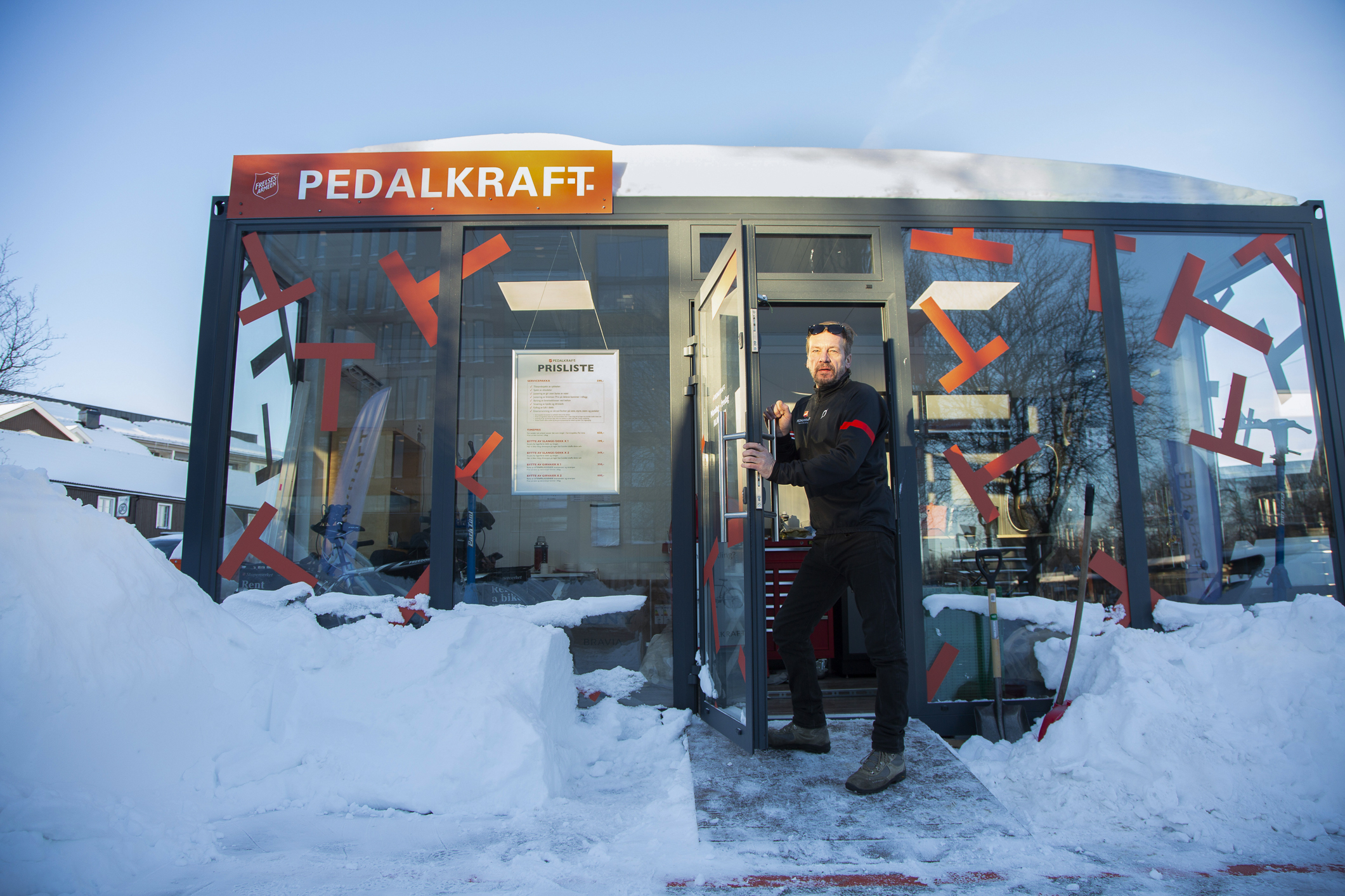 Middelaldrende mann med treningsklær i sort og rødt står utenfor en grønn sykkelbud med glassvegger. Ute der det mye snø