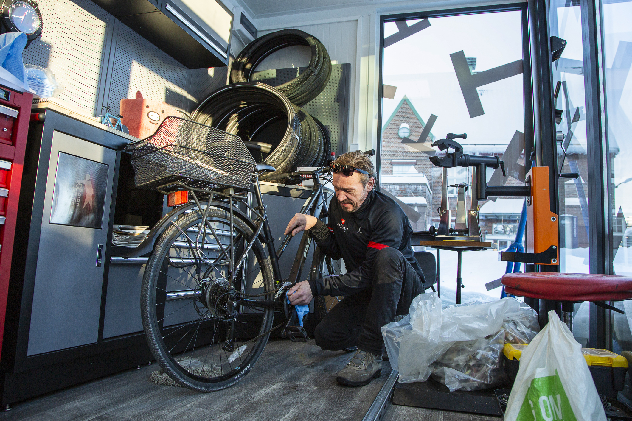 Middelaldrende mann med treningsklær i sort og rødt mekker på en racersykkel i en sykkelbud med glassvegger. Bak ser man sykkeldekk på veggen.