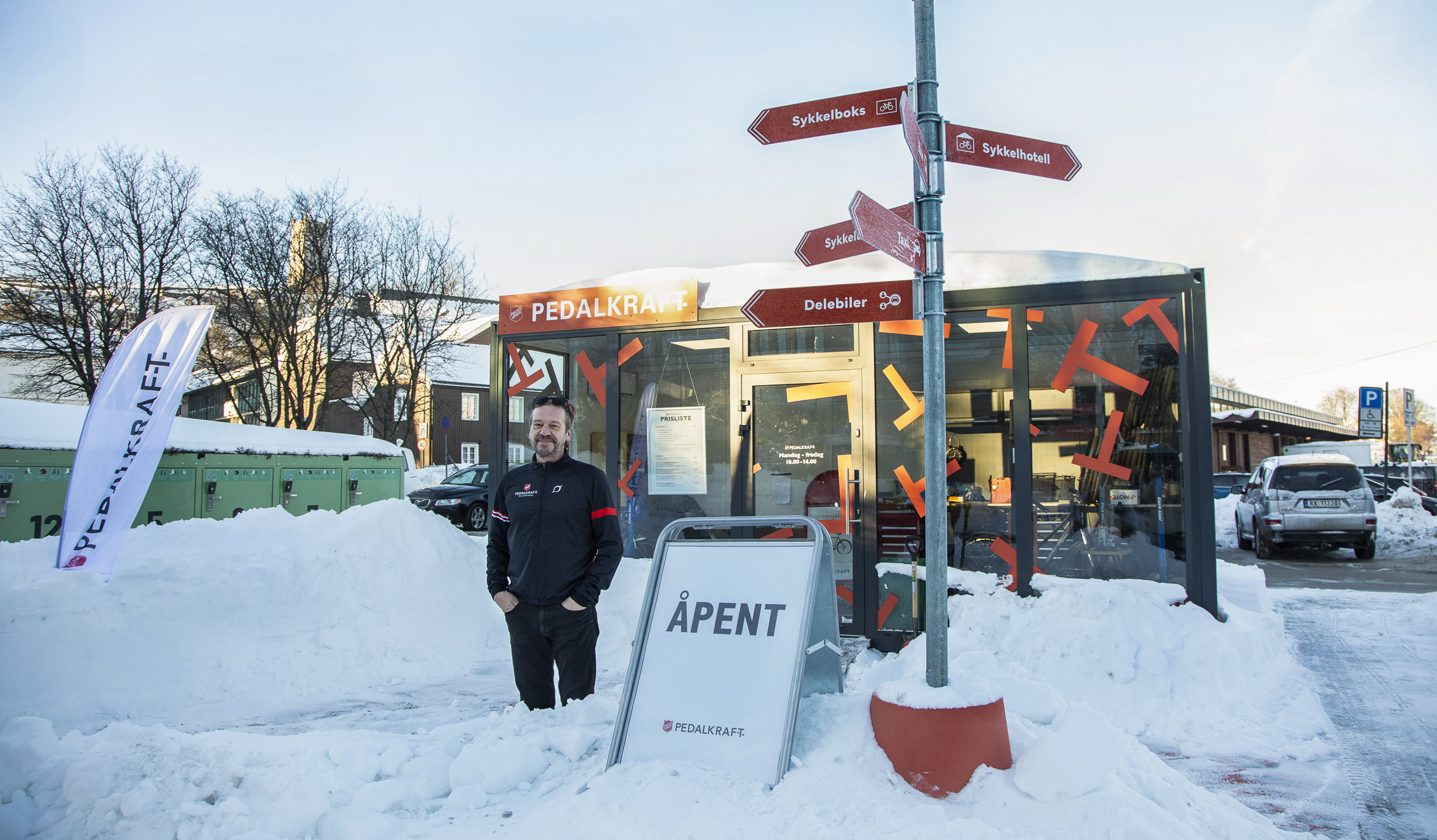 Middelaldrende mann med treningsklær i sort og rødt står utenfor en grønn sykkelbud med glassvegger. Han står ute ved et veiviserskilt. Ute der det mye snø. Det står Pedalkraftopp spå boden og på bakken står det Åpent på et skilt.