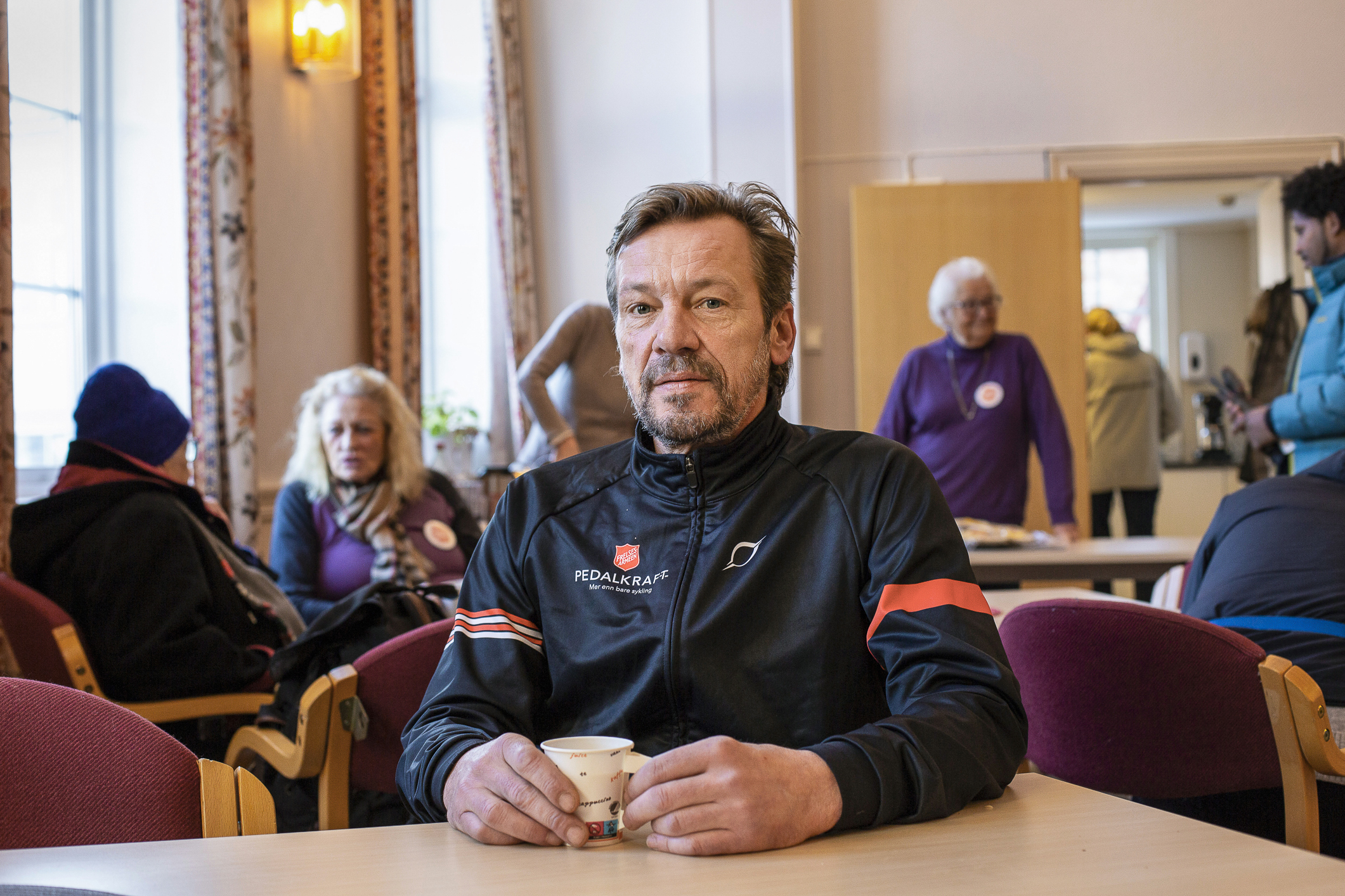 Middelaldrende mann med treningsklær i sort og rødt  sitter ved et bord under matutdelingen i korpset. Han har en pappkopp i hendene.