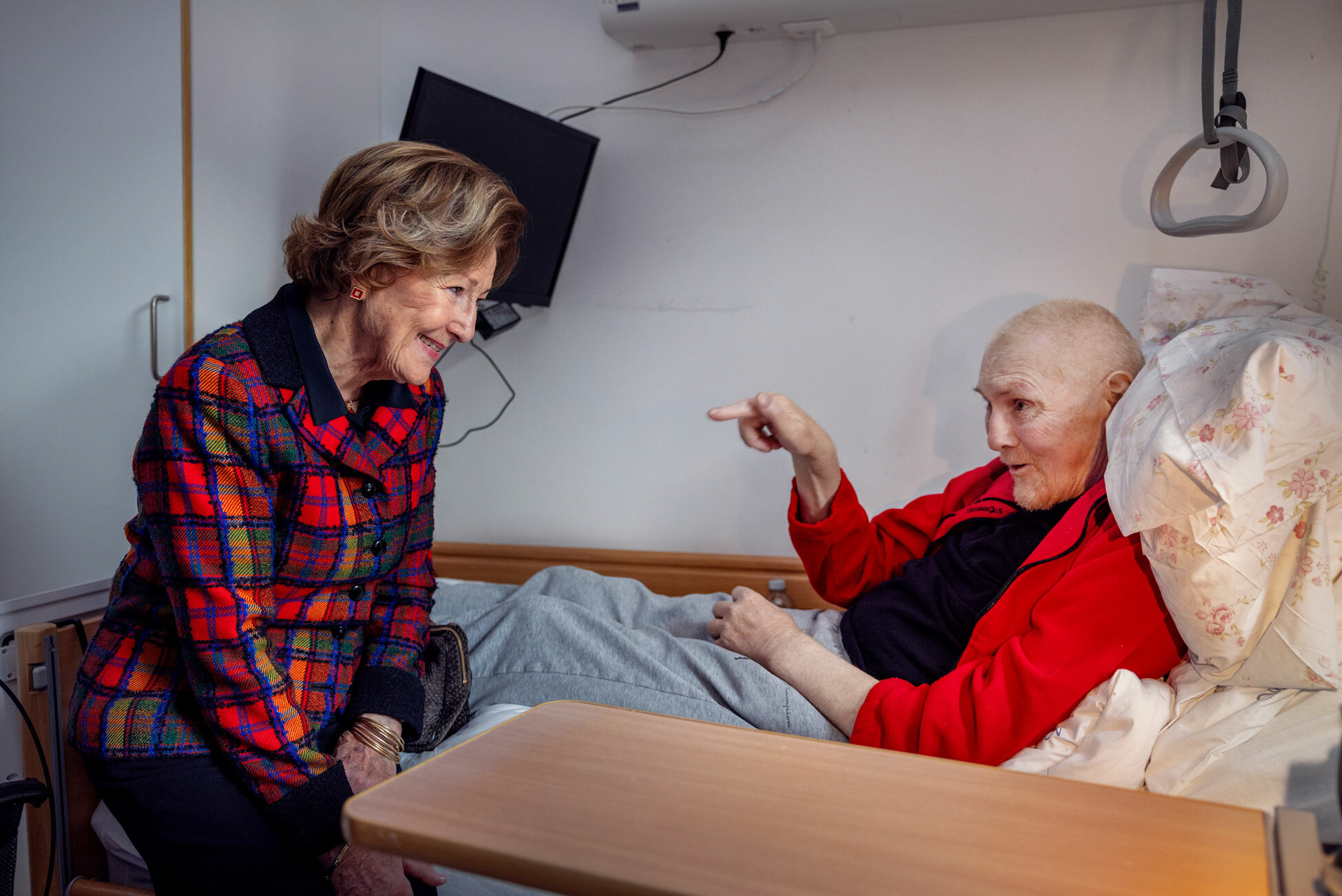 Foto: En kvinne i 80-årene med håret pent frisert sitter på sengekanten til en mann uten hår i 70-årene. Han ligger i en sykeseng med rød flisjakke på seg. Han peker mot damen.