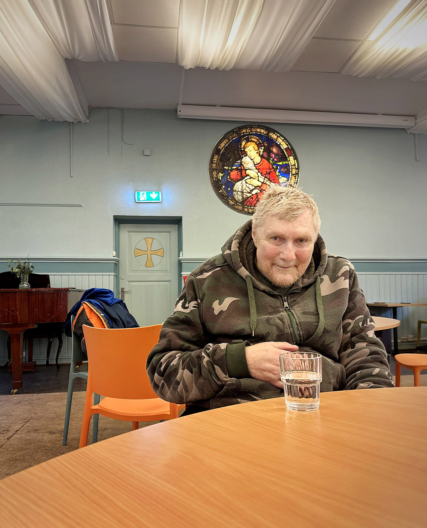 Foto: Eldre mann i kamuflasjejakke sitter ved et bord. I bakgrunnene er det en rundt glassmaleri av Jesus som holder et hvitt lam.