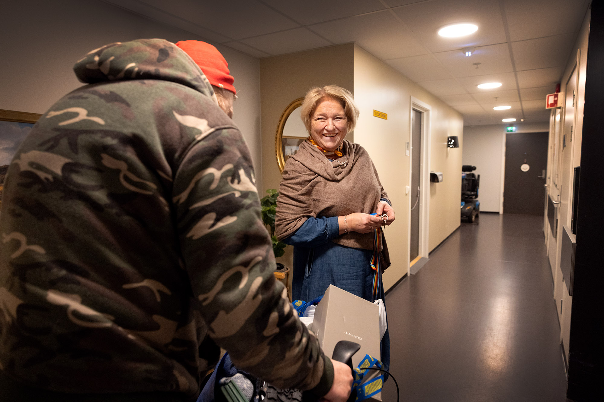 Foto: Kvinne i 50-årene med blondt hår smiler mot en mann i armyjakke som kommer med rullator. På den er en bag fra IKEA.