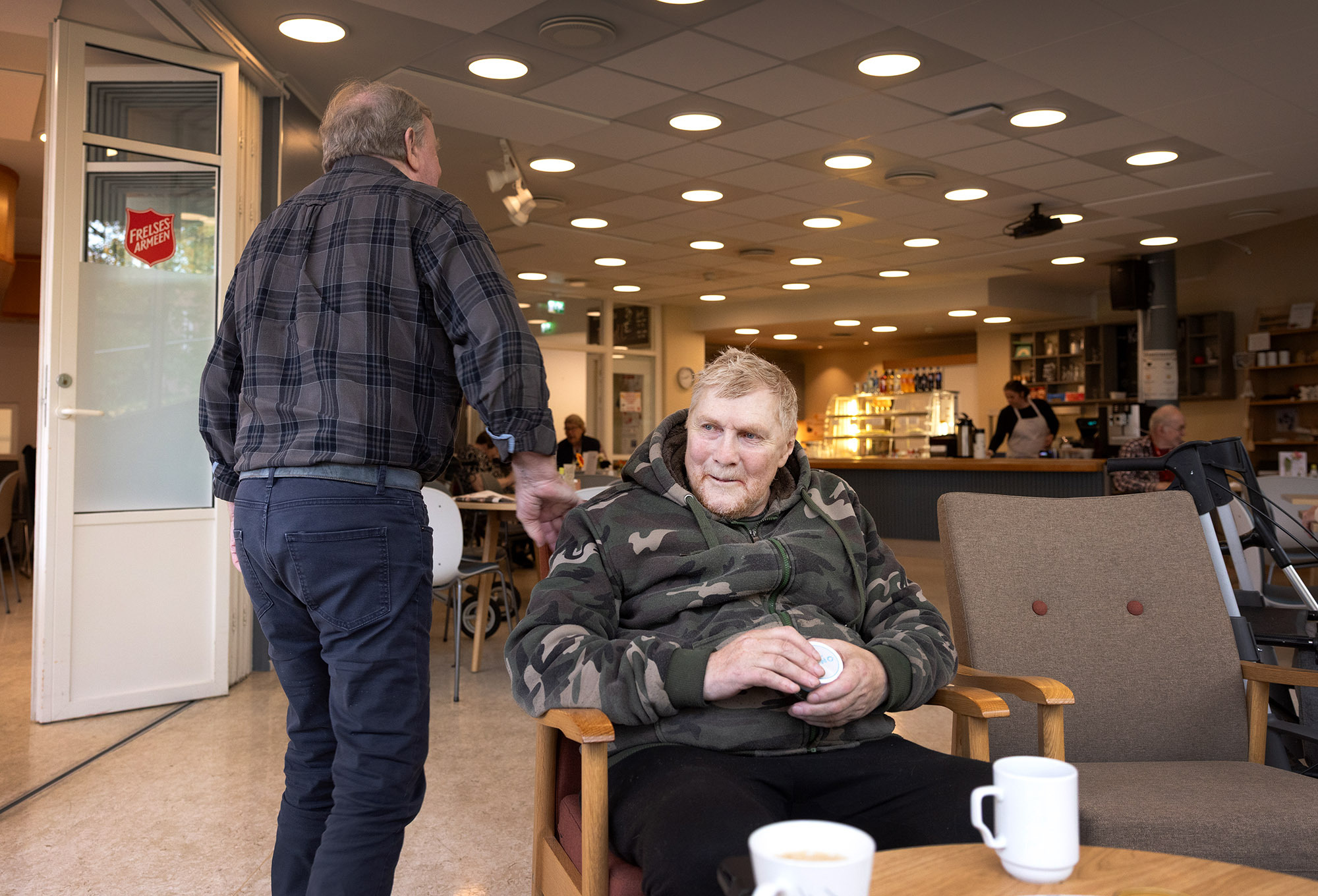 Footo: Mann i armyjakke sitter i en stol. En annen eldre mann går forbi og gir ham et klapp på skulderen.