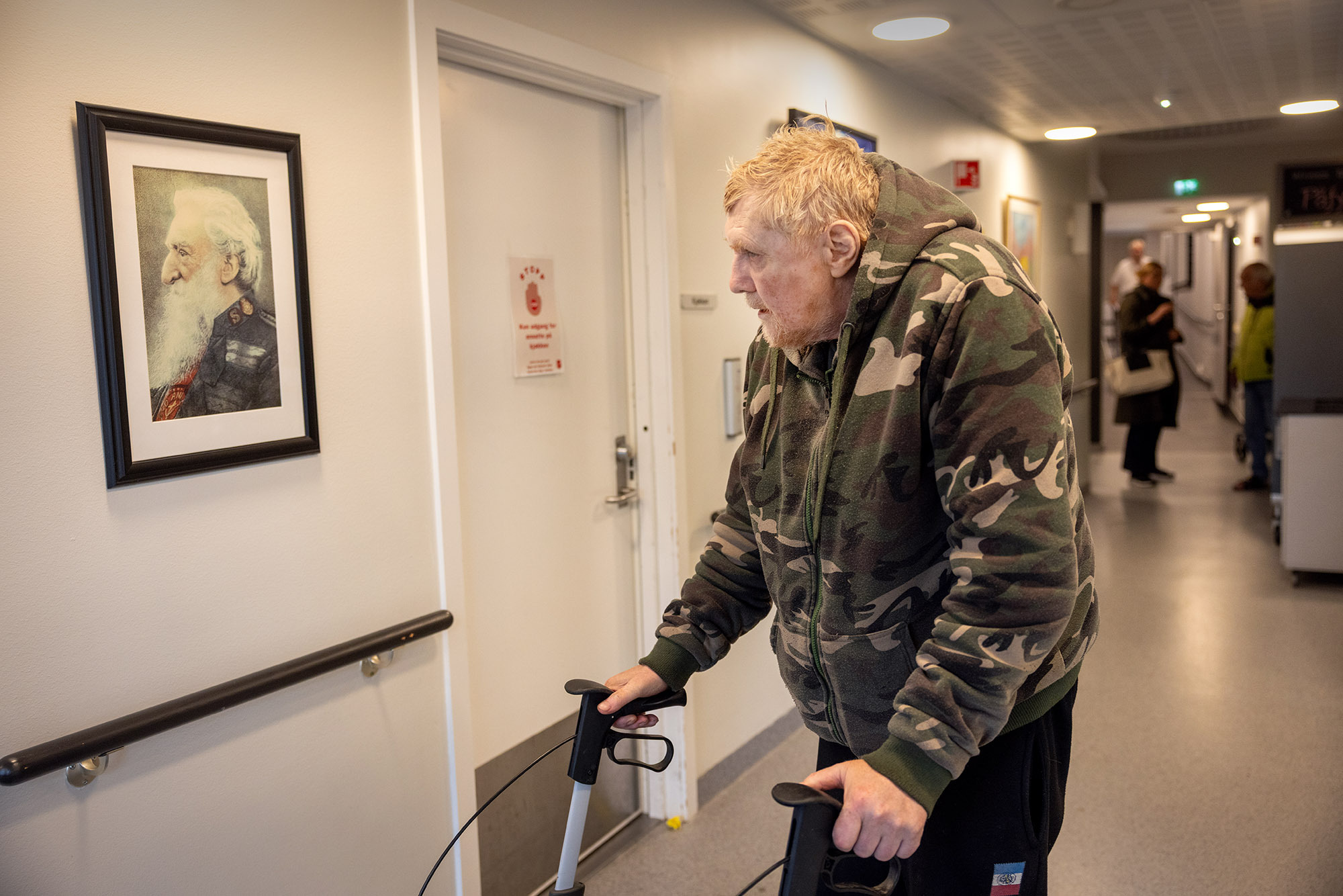 Foto: Mannen i armyjakke ser på et svart-hvitt portrett av en mann med skjegg. Han står i en lang korridor med hvite vegger.