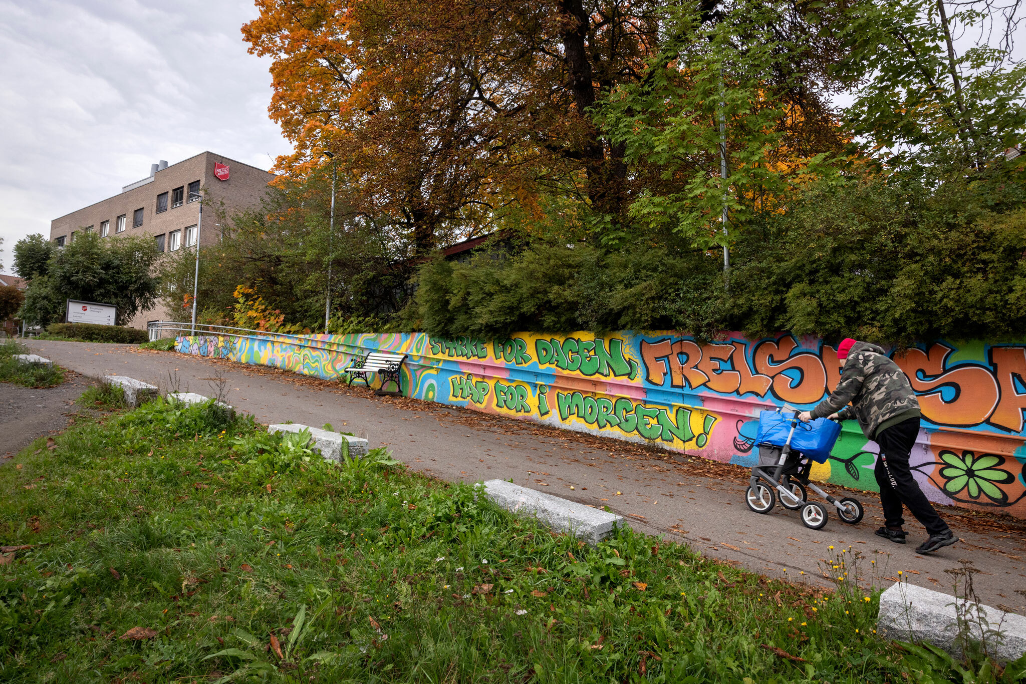 Foto: Mann i armyjakke og rullator går oppover en bratt bakke med fargerik grafitti på muren ved siden av veien. Mursteinsbygget på toppen har en stor, rød logo fra Frelsesarmeen på veggen.