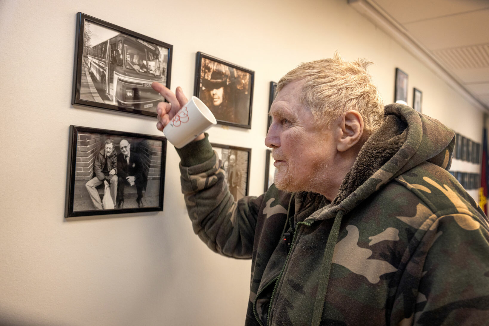 Foto: Den eldre mannen i militærmønstret joggejakke peker på bilder på veggen. I hånden holder han en kopp.