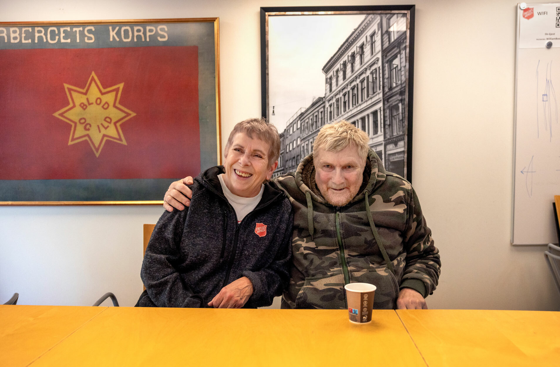 Foto: Den eldre mannen i miltærjakken sitter ved siden av en kvinne i 60-årene. Han har armen godt om hennes skulder. Begge smiler godt. På veggen bak dem henger et gammelt foto av en bygning. Ved siden av på veggen henger et innrammet, rødt flagg fra Frelsesarmeen med en gul stjerne på der det står "blod og ild".