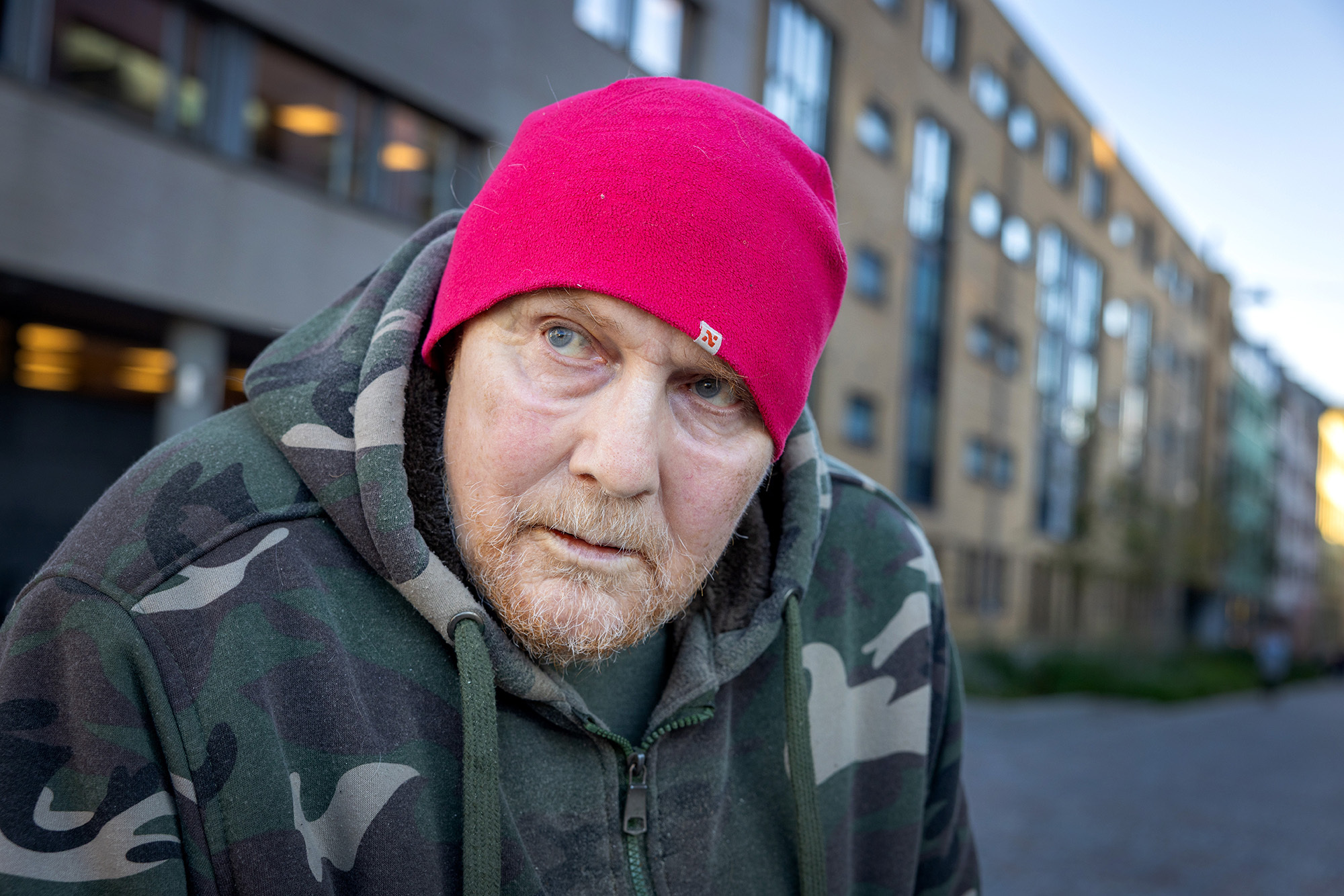 Foto: Nærbilde av eldre mann med rødrosa lue på. Han står ute i en gate og kikker bort. Han har på seg grønn kamuflasjejakke.