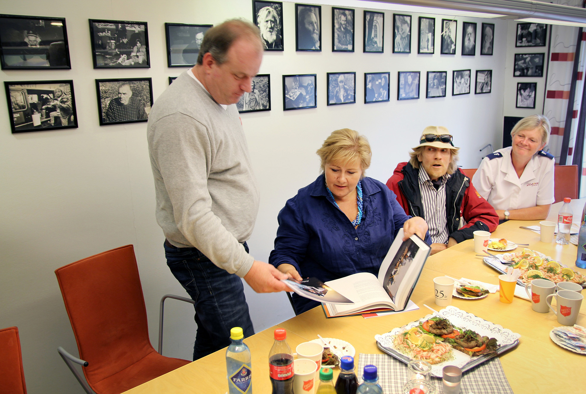 Foto: To kvinner og to menn ved et møtebord i et rom med en rekke bilder på veggen av mennesker. To av dem kikker i et fotoalbum.