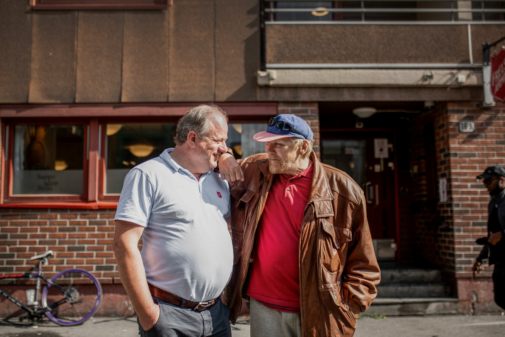 Foto: To menn står tett ved siden av hverandre og snakker ute på gata foran en rød murbygning, som er fyrlyset. Den ene er i 50-årene og den andre i 60-årene.