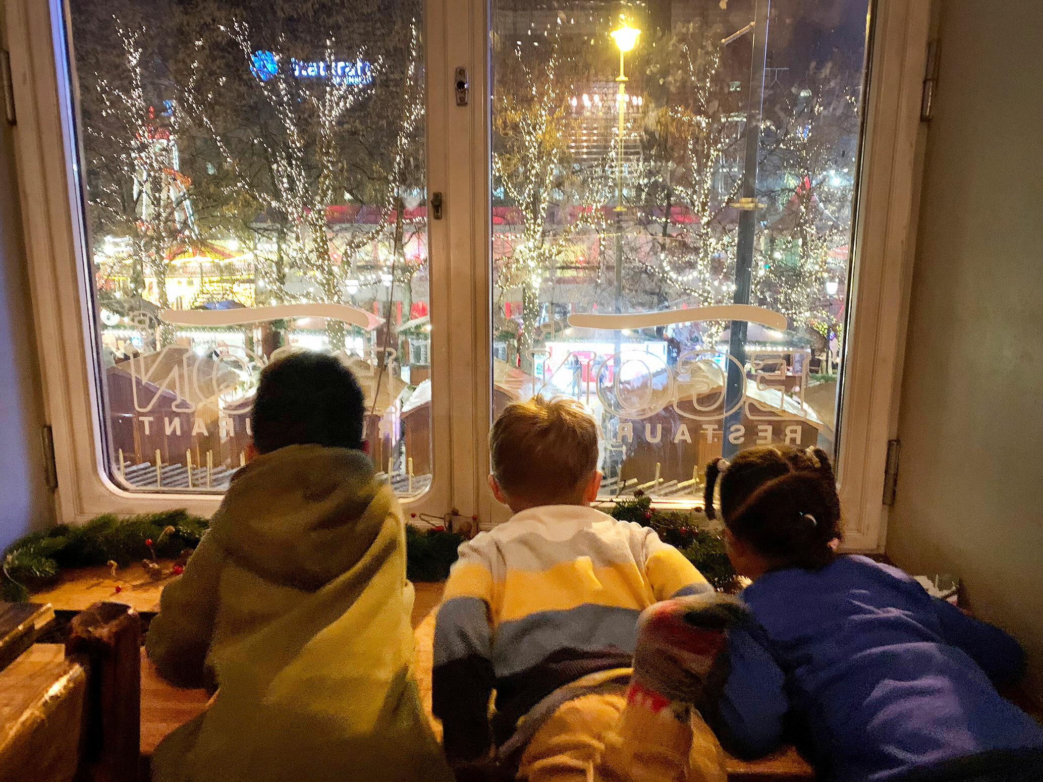 Foto: Tre barn titter ut gjennom et vindu på Egons restaurant på Karl Johan i Oslo. Her ser de ut på isen i Spikersuppa og en julepyntet by.