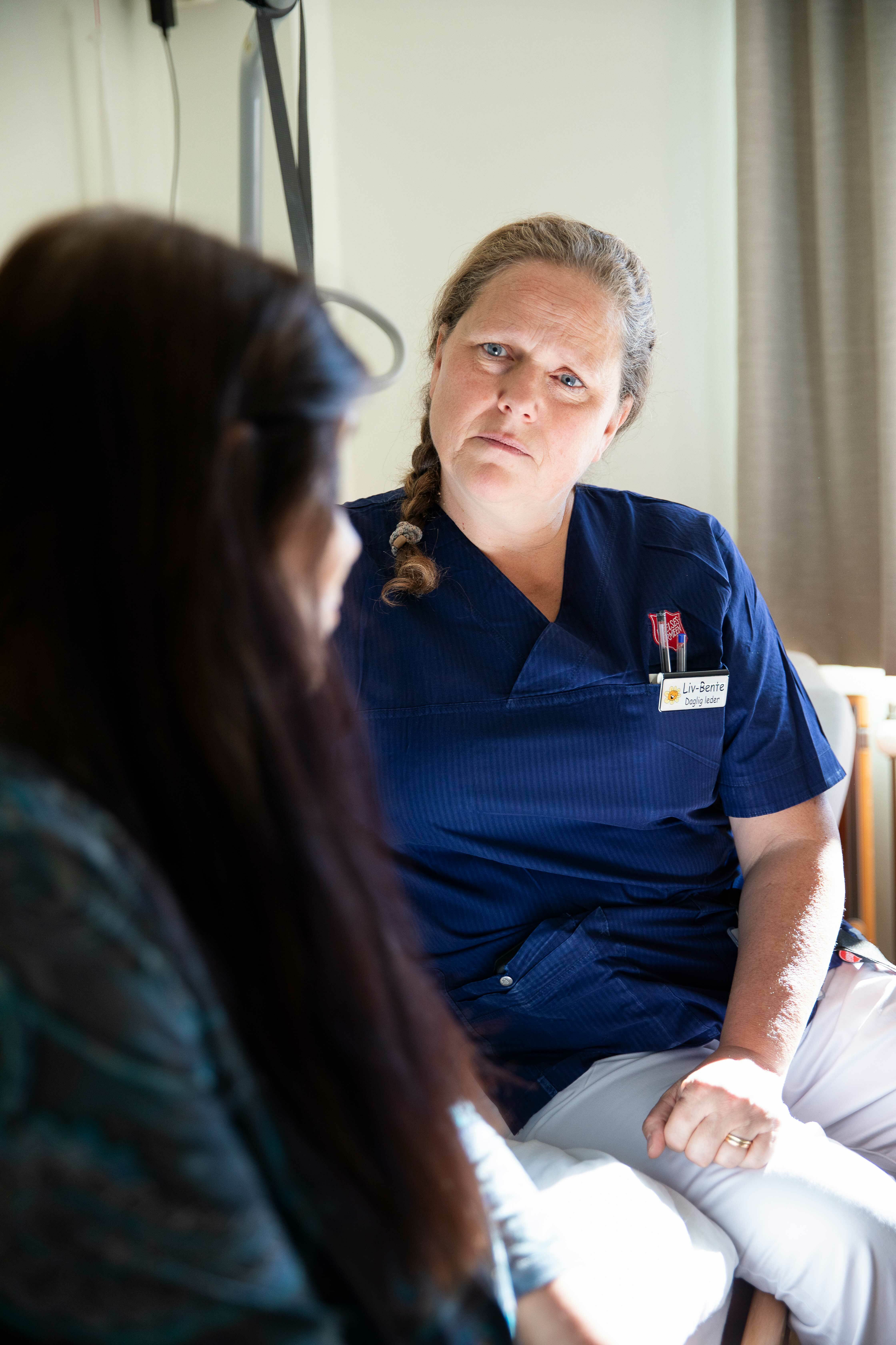 En kvinne i blå t-skjorte sitter og ser på en kvinnelig pasient hun snakker med. Pasienten sitter med siden til og ansiktet er skjult av brunt, langt hår.