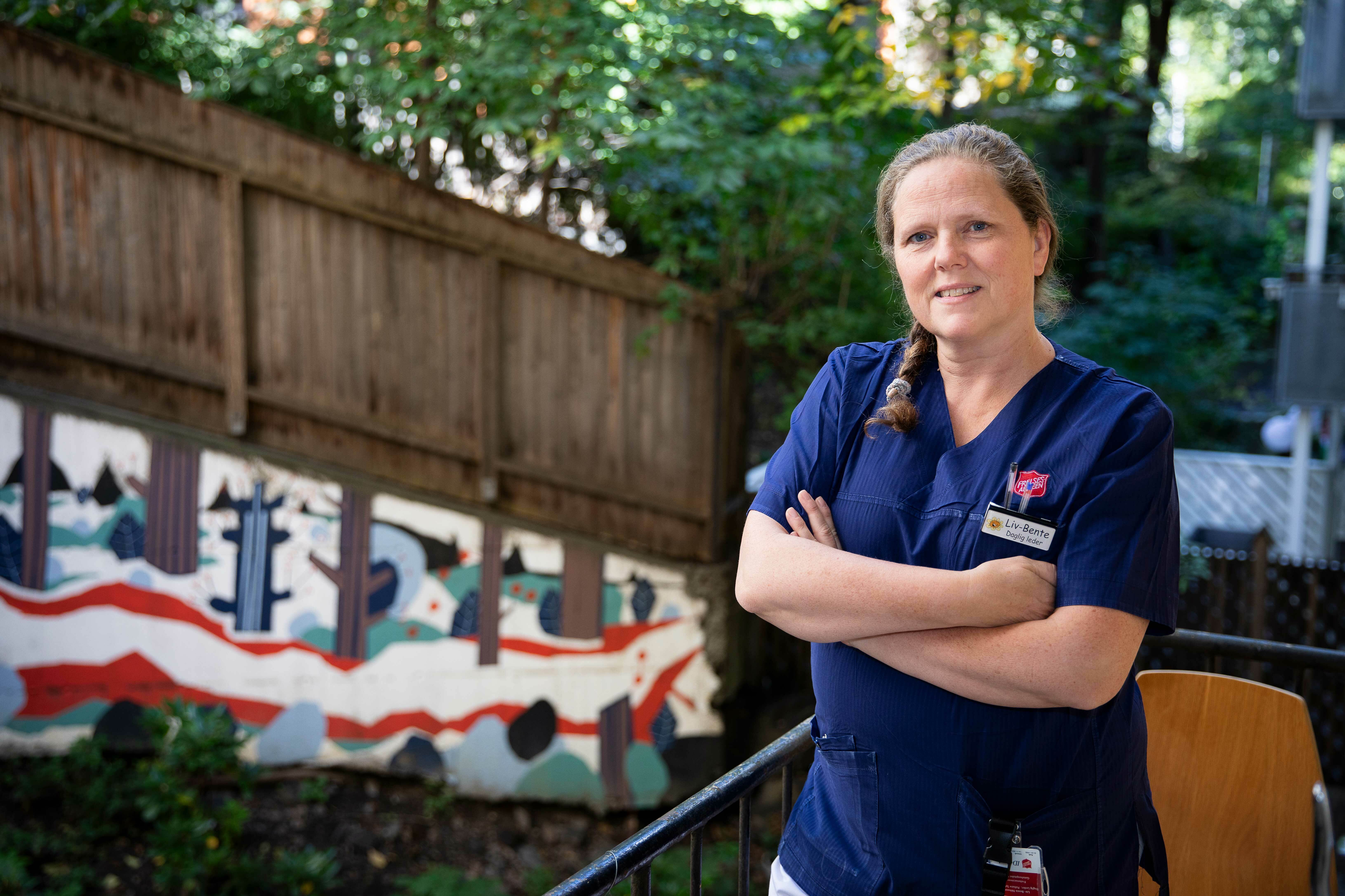 En kvinne med blå t-skjorte står utendørs med armene i kryss foran seg. I bakgrunnen er en dekorert trevegg