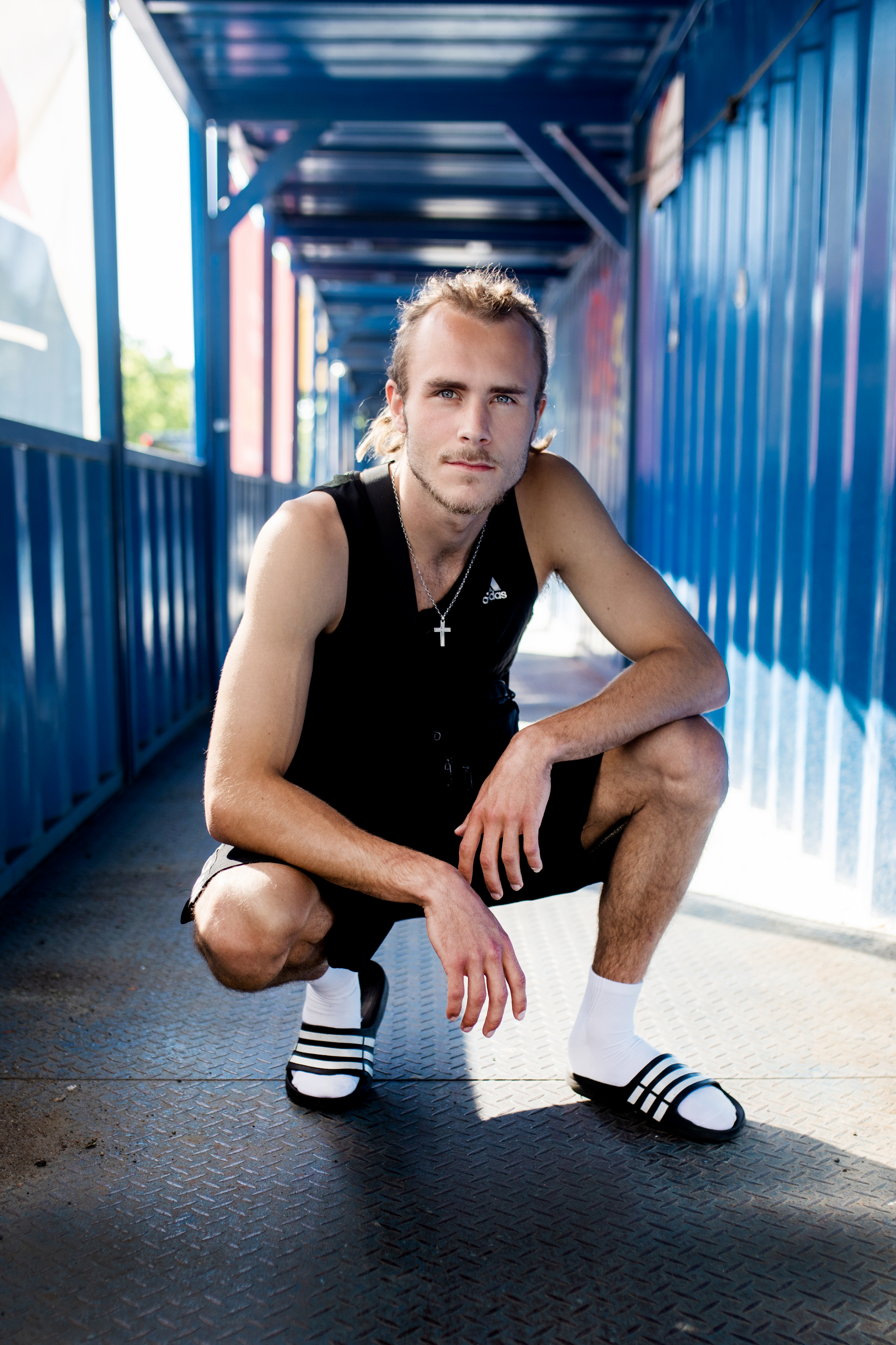 Bildet viser Endre grytting utendørs, han sitter på huk og ser inn i kamera, med en gang med blå metallvegger bak seg.