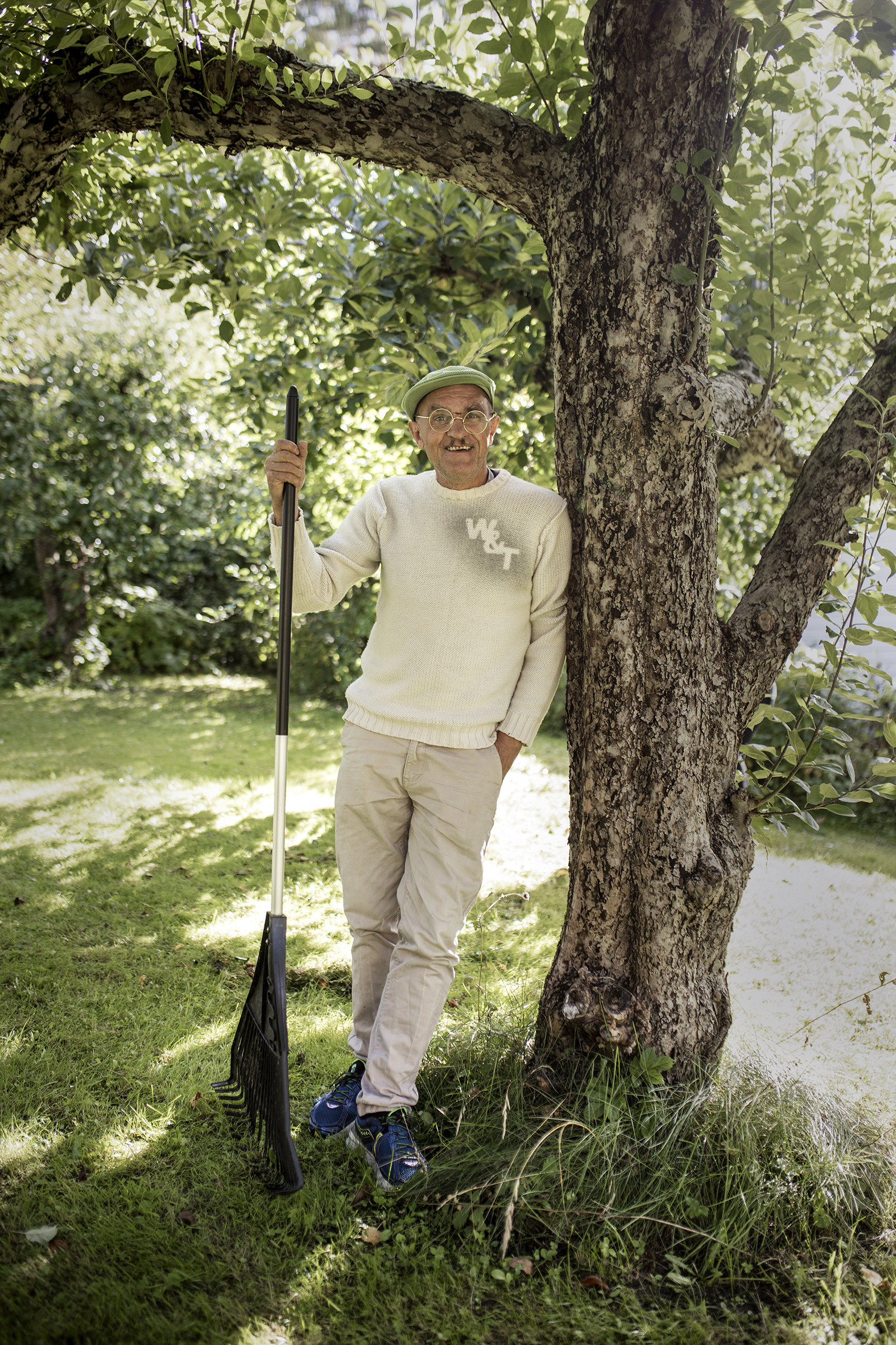 Bildet viser en mann med beige genser, beige bukse og grønn caps, som står og lener seg mot et tre og holder en rake.