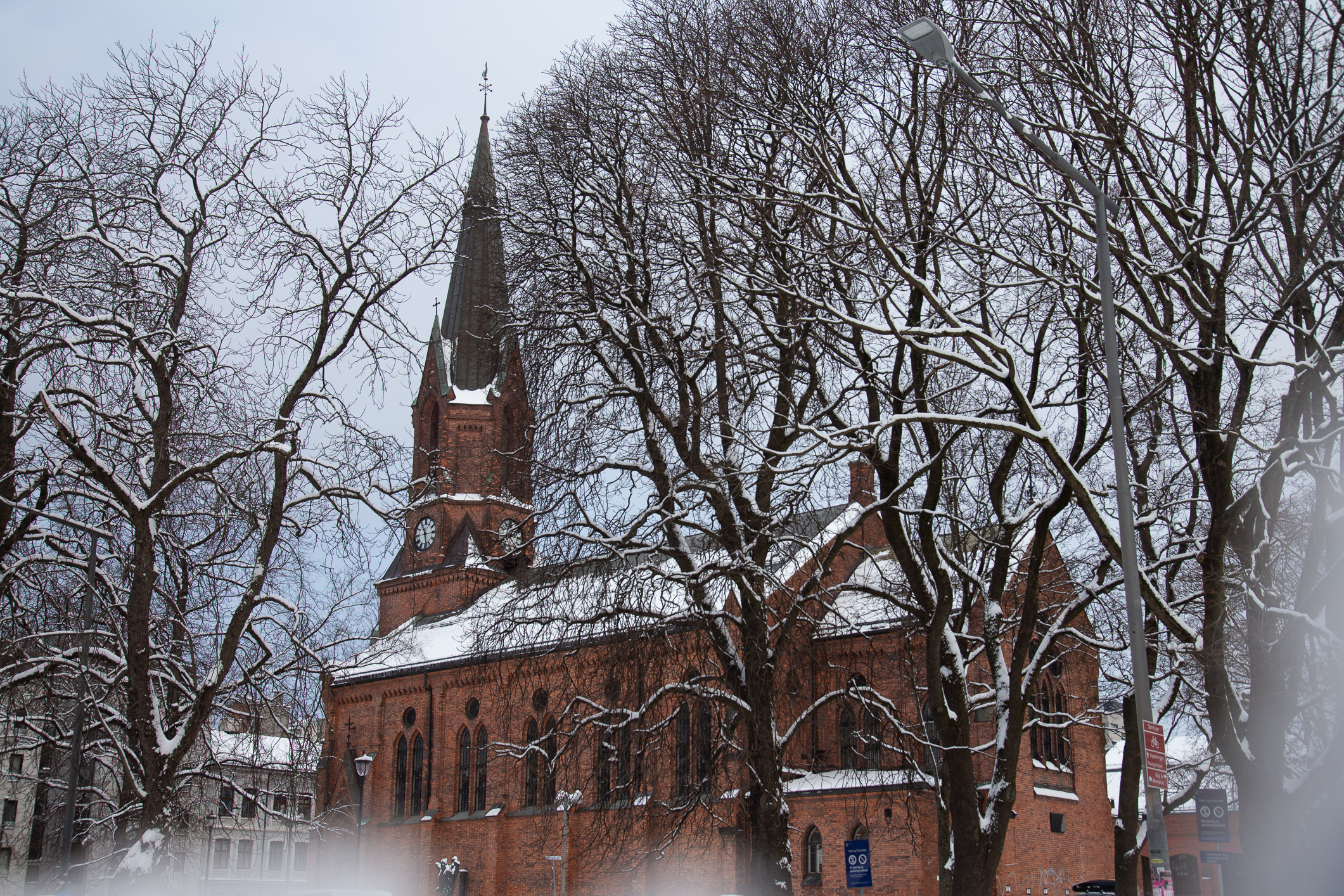 Rød mursteinskirke med spir og trær rundt. Det er vinter og det ligger snø på kirketaket.