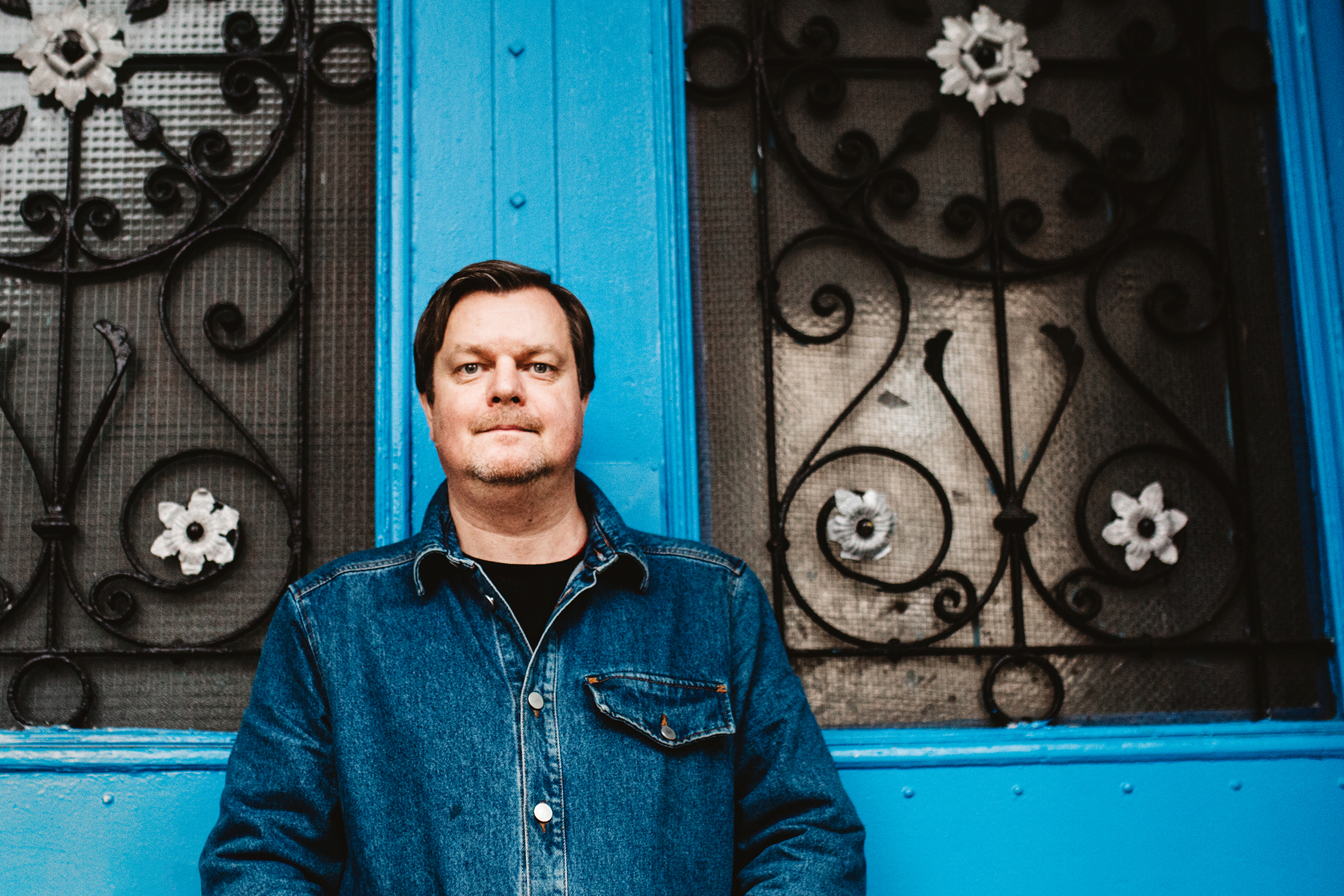 Espen Lervaag står foran en blå dobbeldør med glassruter. Han ser med et nøytralt blikk mot kamera. Han er ikledd en dongerijakke.