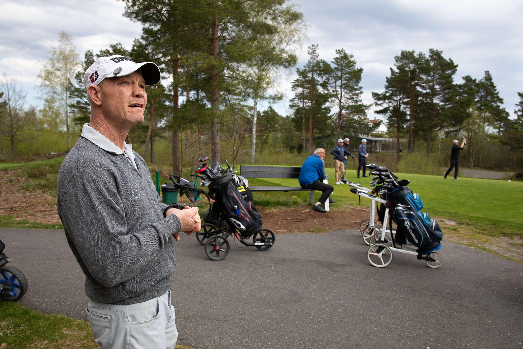 Foto: Mann med hvit skyggelue står i venstre kant av bildet. Rundt ham står en del golfbager parkert og det er mennesker på gresset like ved.