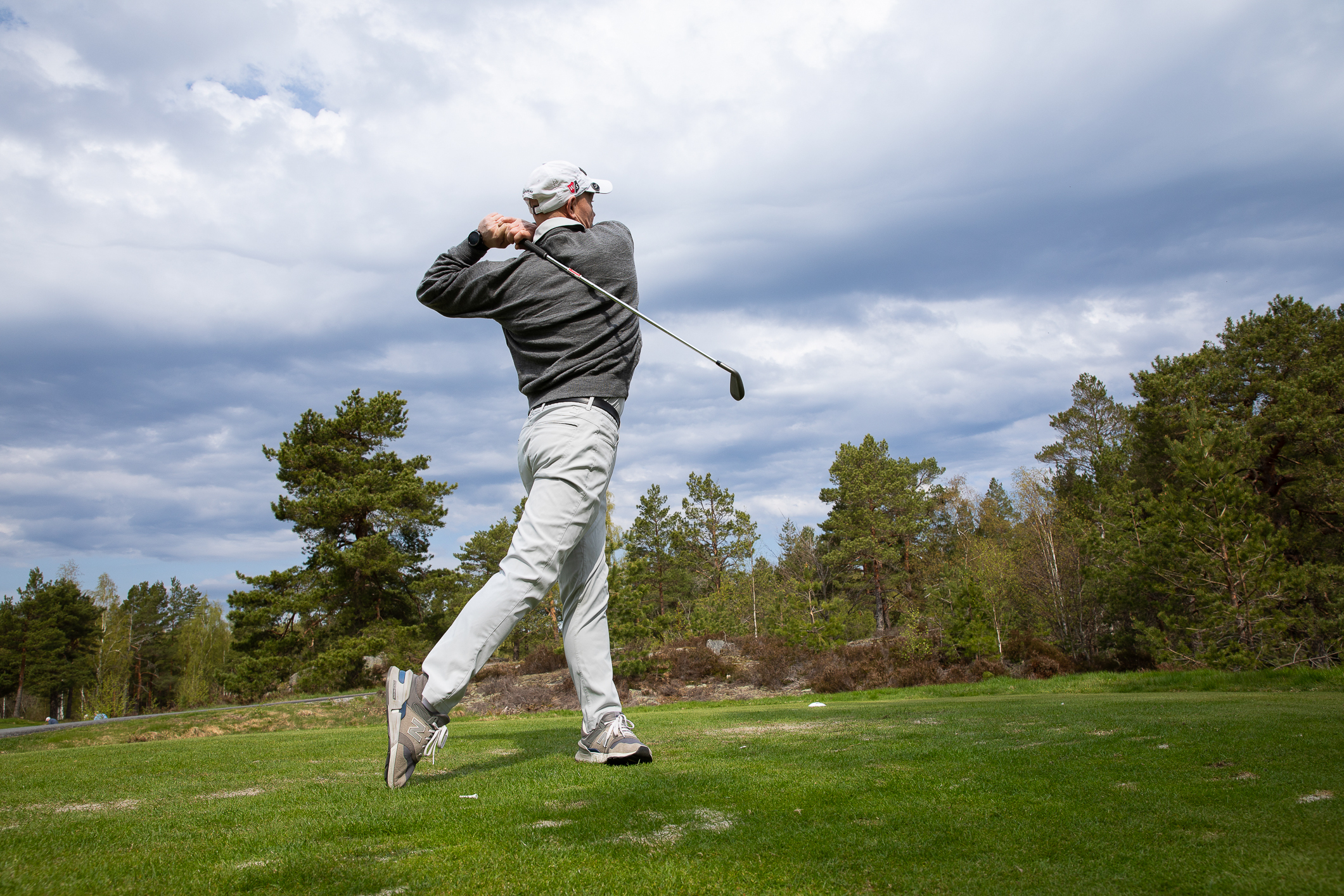 Foto: En mann som akkurat har slått et slag med golfkølla, slik at den er bak på ryggen. Han står på en golfbane med trær i utkanten.