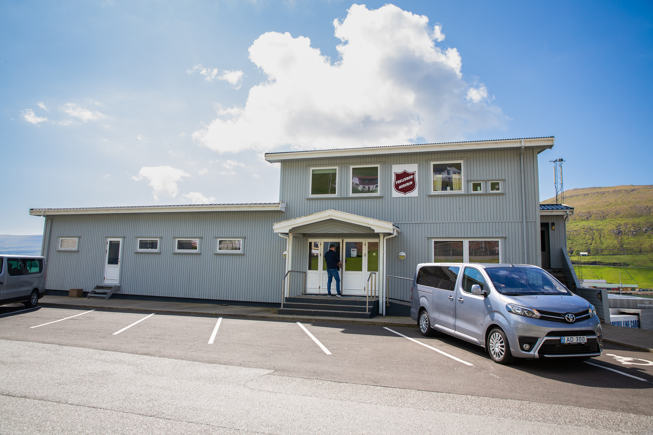 Bilde av grått trebygg med skiltet til Frelsesarmeen på veggen. Utenfor står en varebil parkert og en mann låser seg inn i lokalene.