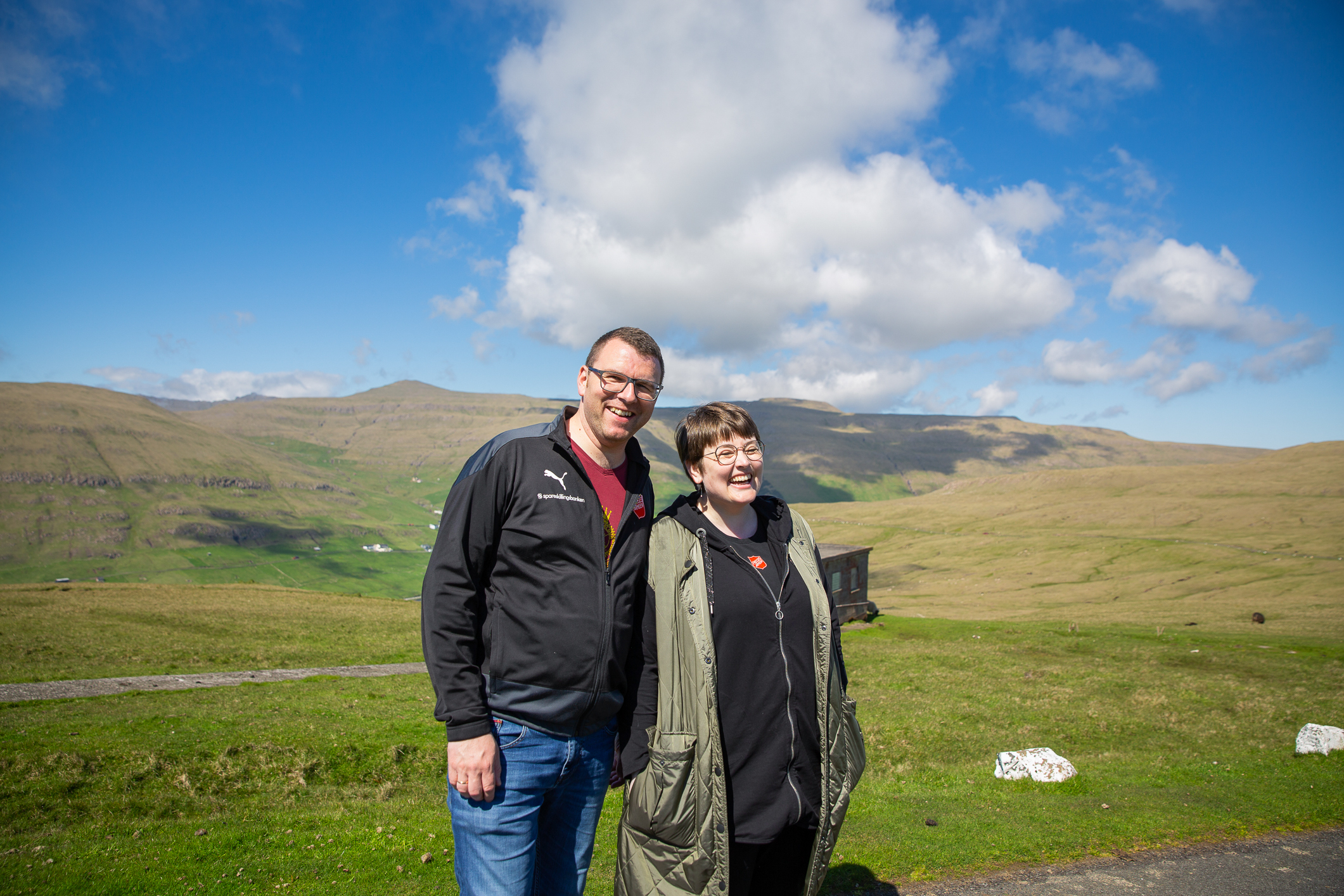 Ektepar som smiler og ler i hverdagsklær. De står på en vidde helt uten trær, kun gress.