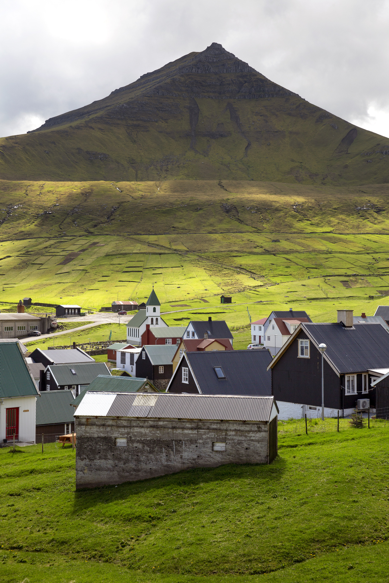 Foto: Et grønt og brunt landskap uten trær med et gjell i bakgrunnen. Det ligger noen få hus nede i en dal.