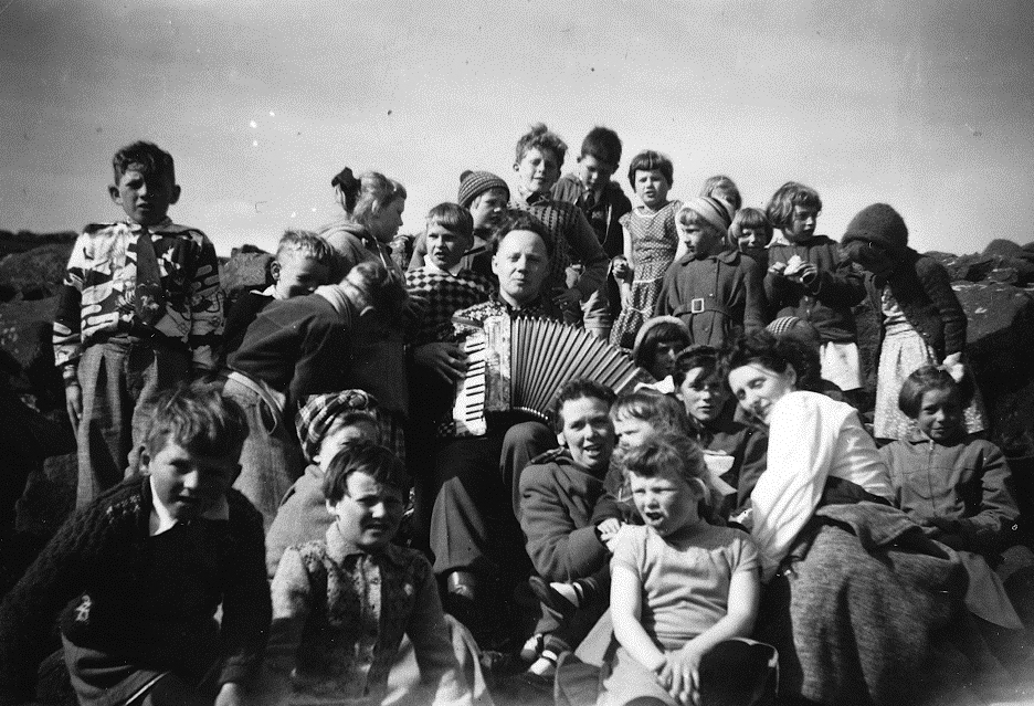 Foto: I svart-hvitt med en mann som spiller trekkspill. Rundt ham sitter mange mennesker – gammel som ung.