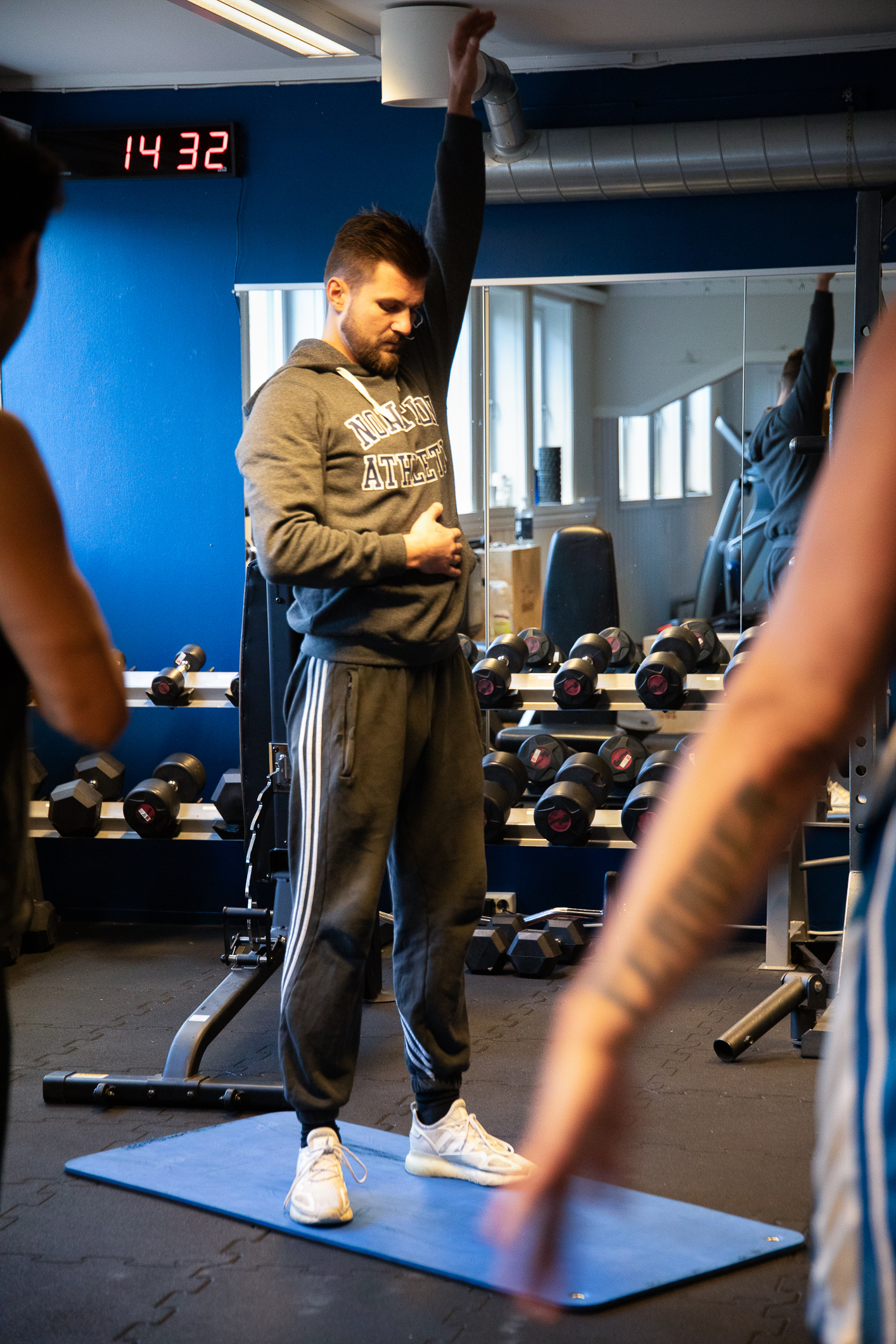 Foto: Mann står i et treningsstudio, på en blå matte. Gjør øvelser ved å strekke venstre arm opp i lufta. I bakgrunnen er det mange håndvekter.