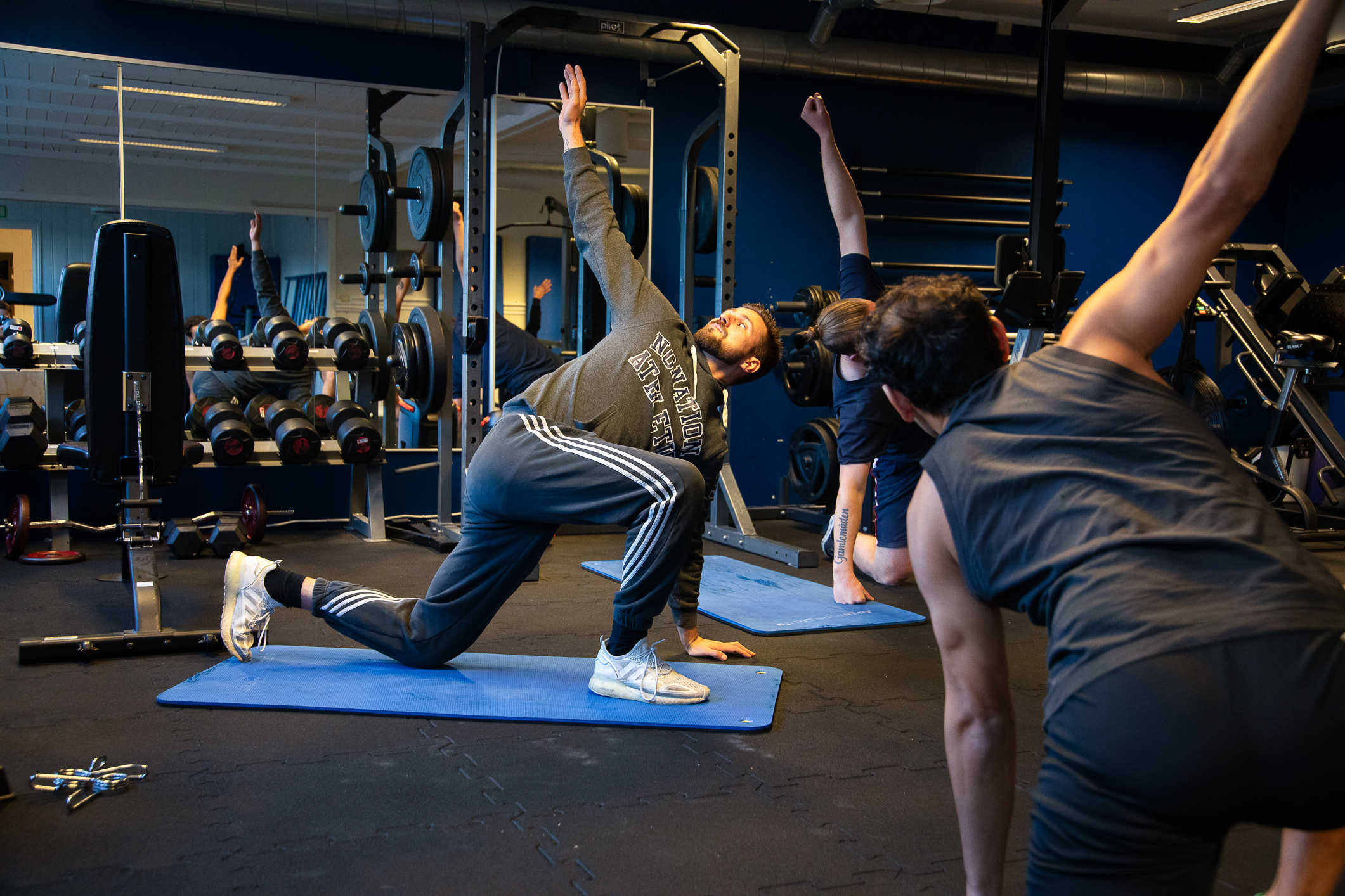 Foto: Tre menn som gjør øvelser på gulvet i et treningsstudio. Vi kan bare se ansiktet til en av dem. De andre to har flere tatoveringer.