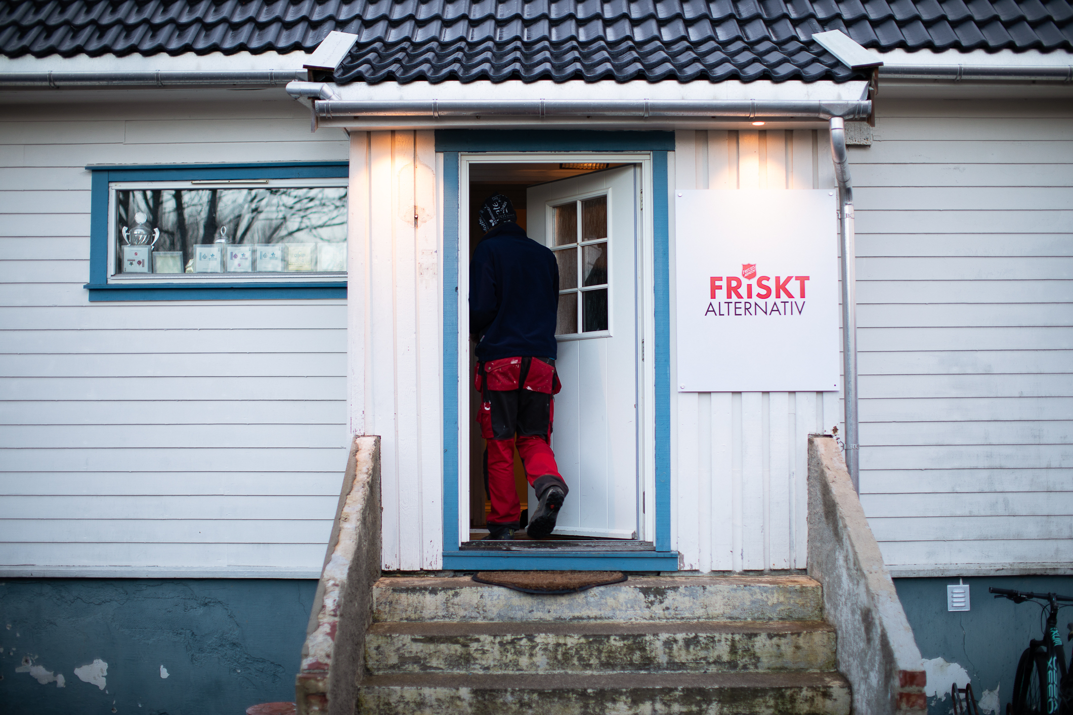 Foto: Ryggen til en person som går inn i et hvitt bygg. Der står Friskt alternativ på et stort skilt utenfor døra med Frelsesarmeens logo på.
