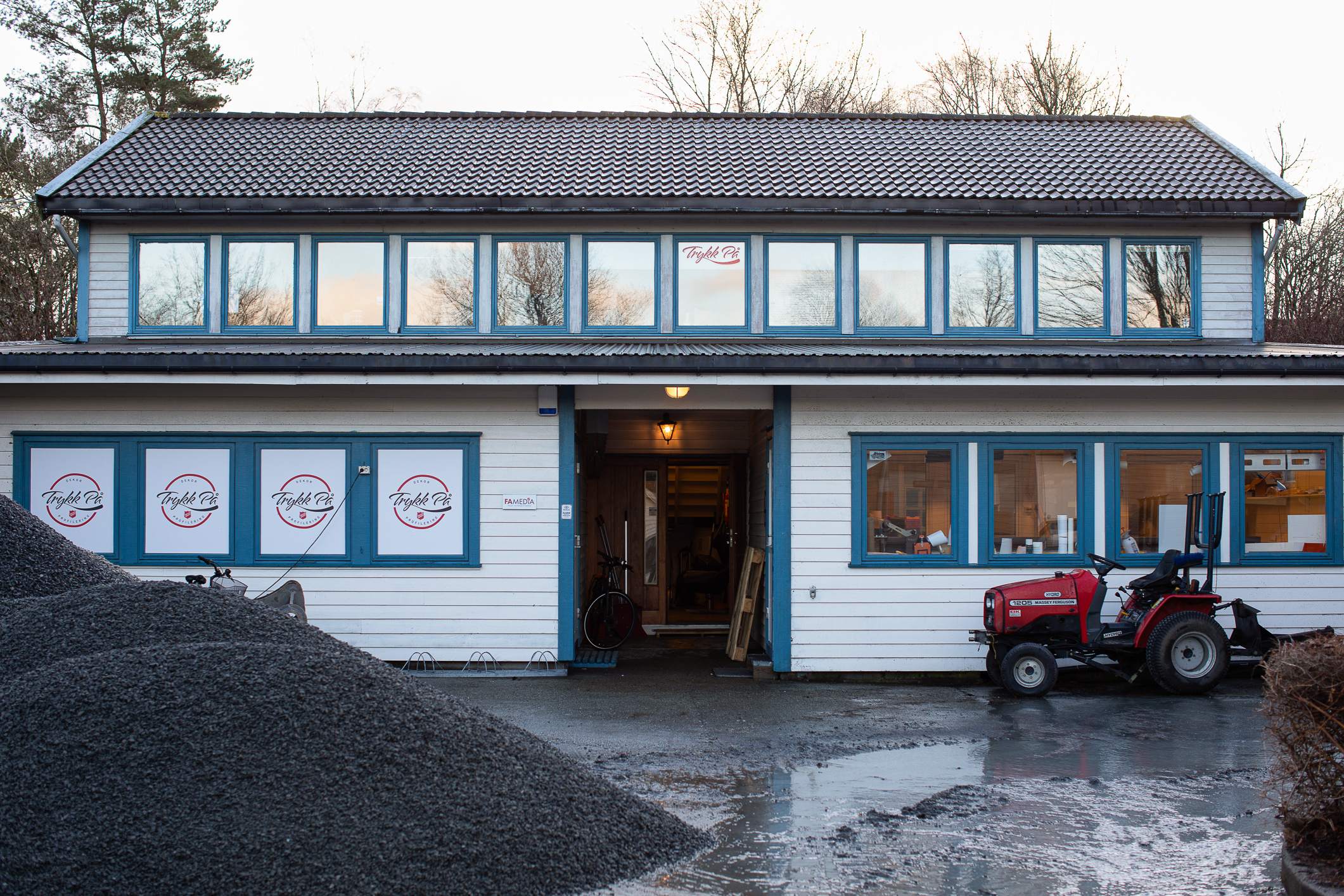 Foto: Hvitt, lavt hus med blå vinduskarmer. I vinduene står logoen Trykk på. Foran bygget står en liten rød traktor parkert.