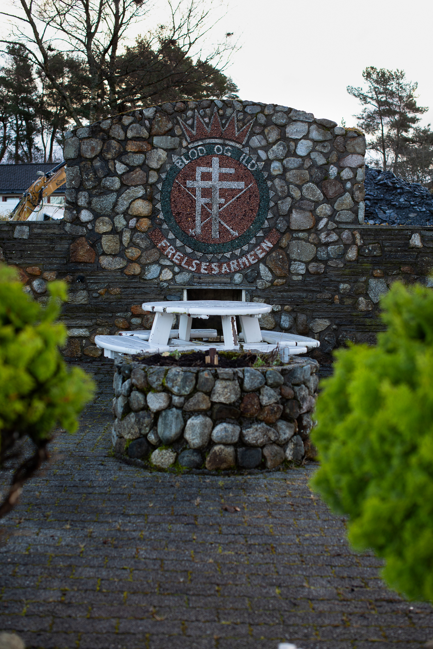 Foto: Frelsesarmeens logo bygget inn i en høy mur i rullestein. Foran er det murt opp en rund brønn i samme type stein med en rundt, hvit sittebenk over.