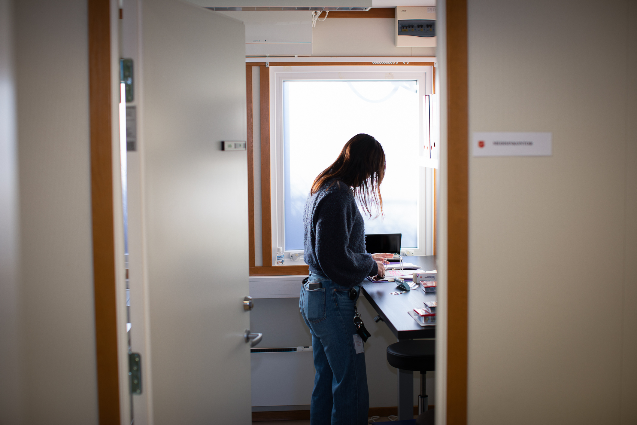 Foto: Kvinne står alene på kjøkkenet. Det mørke håret dekker ansiktet der hun står med sin høre side vendte mot kamera. I bakgrunnen er det et stort vindu.